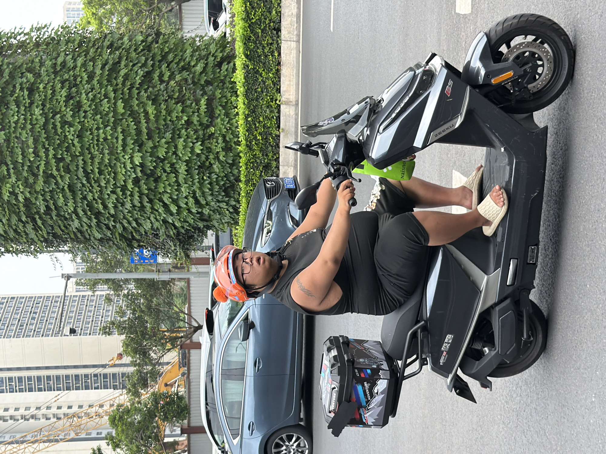This image captures an outdoor urban scene in the Chongqing High-tech Zone, China, during daylight hours. A single individual is depicted riding a dark grey electric scooter on a paved road, facing slightly towards the right.
The rider is an adult male with a heavy build, wearing a black sleeveless top and black shorts. He has a tattoo on his upper left arm. He wears black-rimmed glasses and an orange helmet adorned with two small, fluffy orange protrusions resembling ears. His mouth is slightly open, and he is looking towards the right side of the frame. He wears light-colored (possibly beige or off-white) slide sandals on his feet, which are positioned on the scooter's footboard. His hands are on the handlebars, suggesting the scooter is in operation.
The scooter, identified by "ZEEKU" and "GS" branding on its front fairing, features a clear windscreen and disc brakes on the front wheel. A light green shopping bag, partially displaying "Naisno," hangs from the handlebars. Attached to the rear of the scooter is a black cargo box adorned with white, blue, red, and grey graphics depicting mountains and the word "ADVENTURE." A black strap or flap is visible on the left side of this box.
The background reveals a multi-layered urban environment. To the far left, several tall, light-colored buildings with numerous windows rise into the sky. A large yellow construction crane is visible among these structures, with a portion of a yellow excavator also seen at ground level. In the mid-ground, a dark blue Mazda sedan is parked or moving slowly behind the scooter rider, with another dark vehicle partially visible behind it. A low, trimmed green hedge runs horizontally behind these cars. A brown fence or wall with a white top edge is also visible further back. To the right, a large, cylindrical mass is densely covered in green ivy. Adjacent to this, a grey street light pole supports what appears to be a security camera and a partially obscured blue road sign. The road surface is asphalt, with white painted lane markings visible beneath the scooter's front wheel.