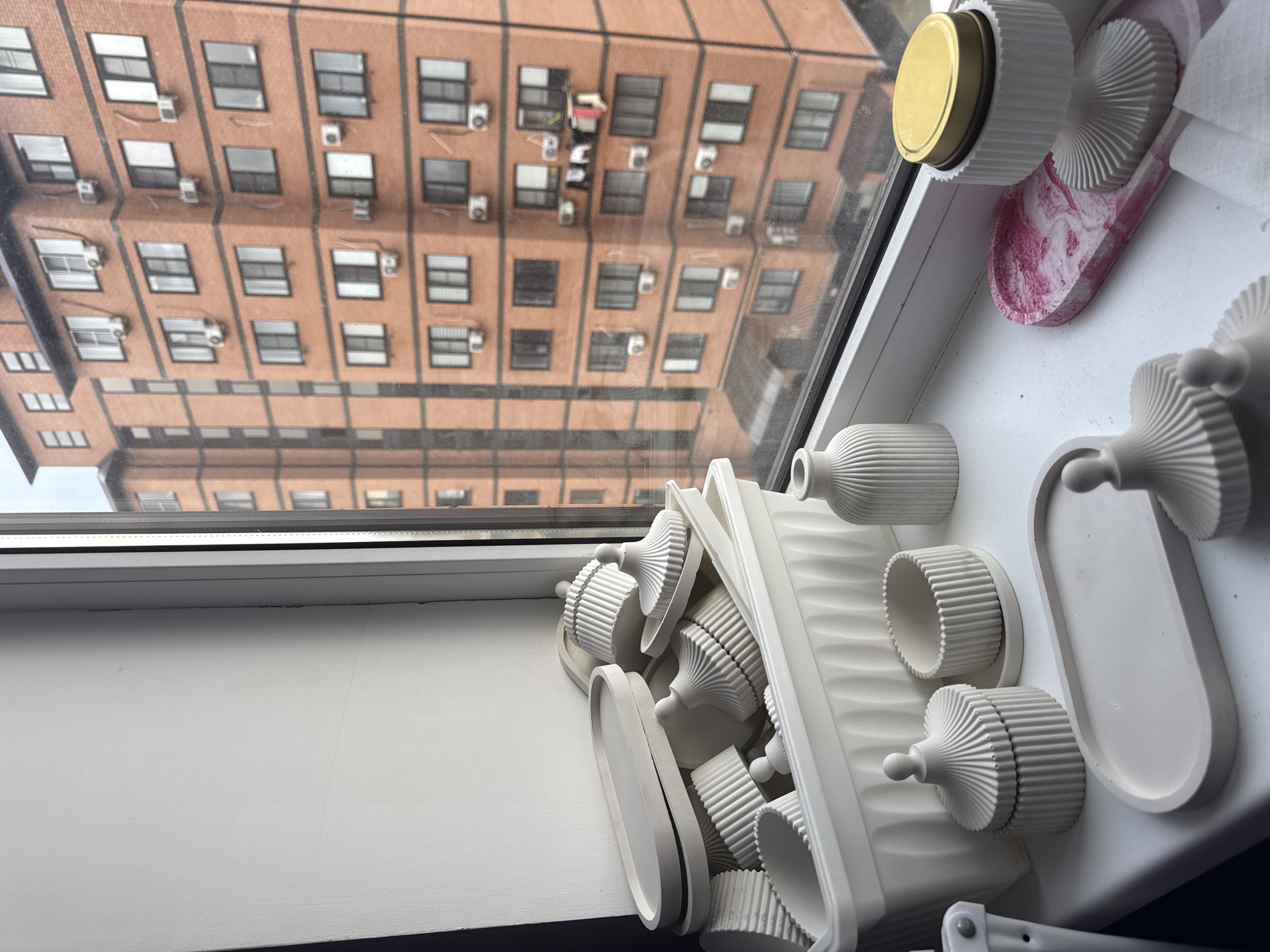 A high-angle shot depicts a window view, showcasing both interior objects and the urban exterior of the city, Unknown, Unknown. Inside, a white windowsill hosts a collection of decorative plaster objects: cylindrical containers, plates, and a small container with a gold lid, along with a pink and white marbled item. Outside, a brick building is visible through the window, featuring numerous windows with dark frames and several wall-mounted air conditioning units. Some clothing is hanging from one of the windows. The overall scene suggests a contemporary living space within an urban environment.