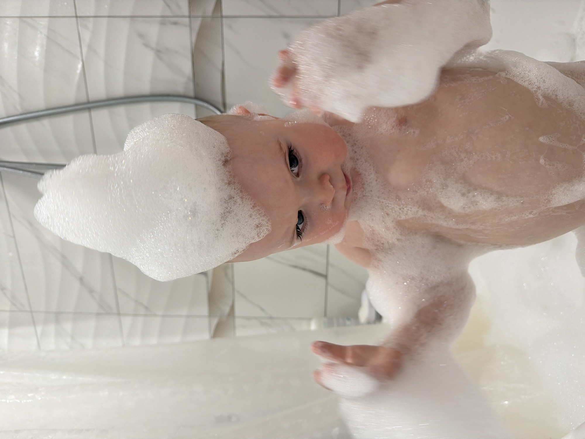 A baby is submerged in a bathtub filled with white bubbles. The baby has bubbles piled on its head and body, with only a portion of its face visible. The backdrop consists of white wall tiles, a shower hose, and a partially obscured shower curtain. Location information (Votkinsk, Russia) is not discernible from the image.
