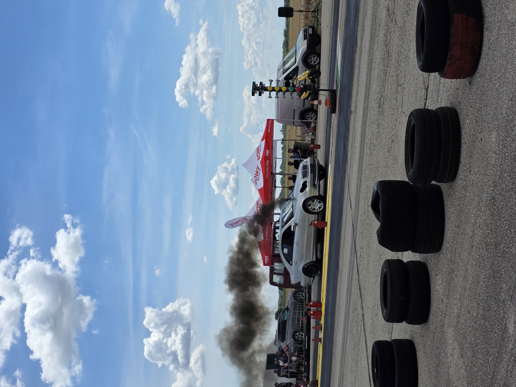 The image depicts a drag racing event at the Akatsievo airfield in Bulgaria. Two cars are positioned on the asphalt track, with the car on the right emitting a large plume of black exhaust smoke, indicating it is either accelerating or has just done so. In the background, several tents with "ROWE MOTOR OIL" branding are set up, suggesting sponsorship. A red van is parked behind a barrier, and a silver van is visible further down the track. A yellow motorbike is parked near a Christmas tree light system used for starting the races. Several tires are stacked on the side of the track in the foreground. The sky is blue with scattered white clouds. The overall scene captures a moment of intense automotive activity during a daytime event.