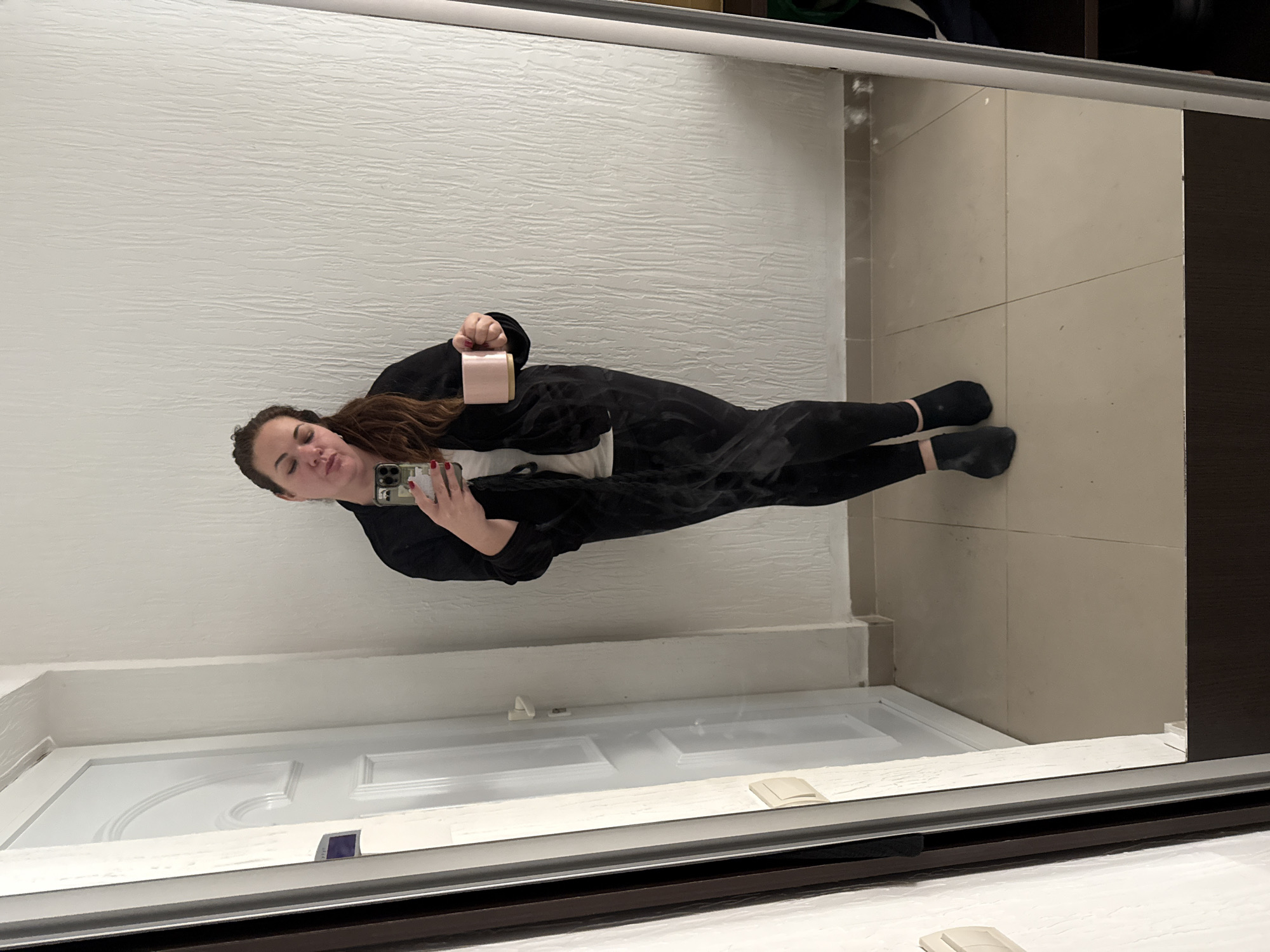 A mirror selfie shows a woman standing in a hallway of what appears to be a residential interior. She is holding a phone in her right hand, taking the picture, and a small, light pink cup in her left hand. She is wearing black clothing and black socks, her hair is brown. Behind her is a textured white wall and a closed, white door. The flooring is light-colored tile. The location is indicated to be Bijela, Montenegro.