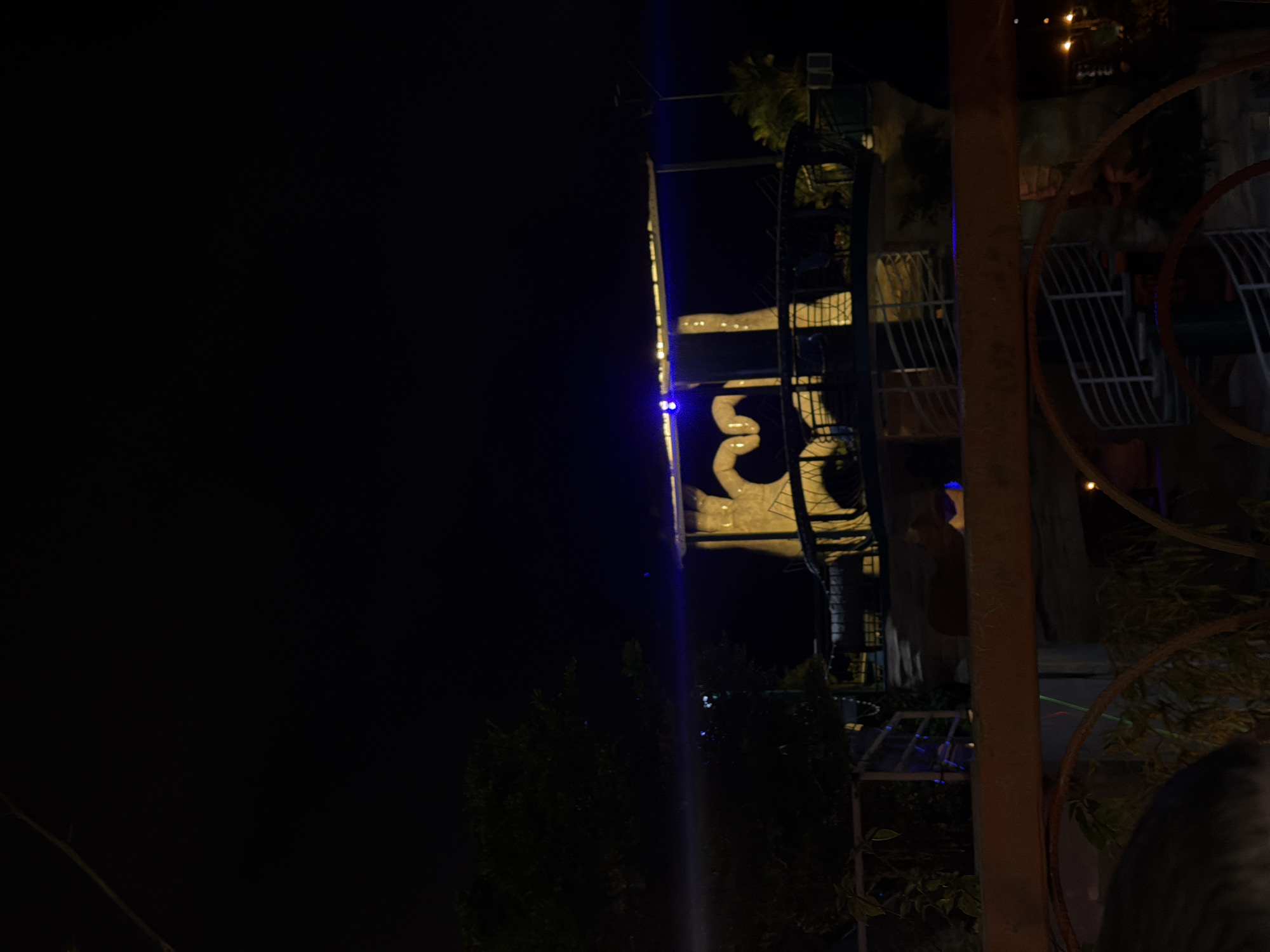 A nighttime outdoor scene in Nam Nha Trang Ward, Vietnam, features a structure with a large heart shape illuminated by lights. The structure appears to be an elevated platform or building, possibly part of a restaurant or entertainment venue, with a dark, open sky above. Trees and foliage are visible on the left side of the image, and lighting fixtures, including what appears to be a street lamp, are present in the background. The foreground shows a railing and what might be the top of a vehicle or another structure, obscuring the lower portion of the scene. Blue lights are prominent on the main structure, casting a glow.