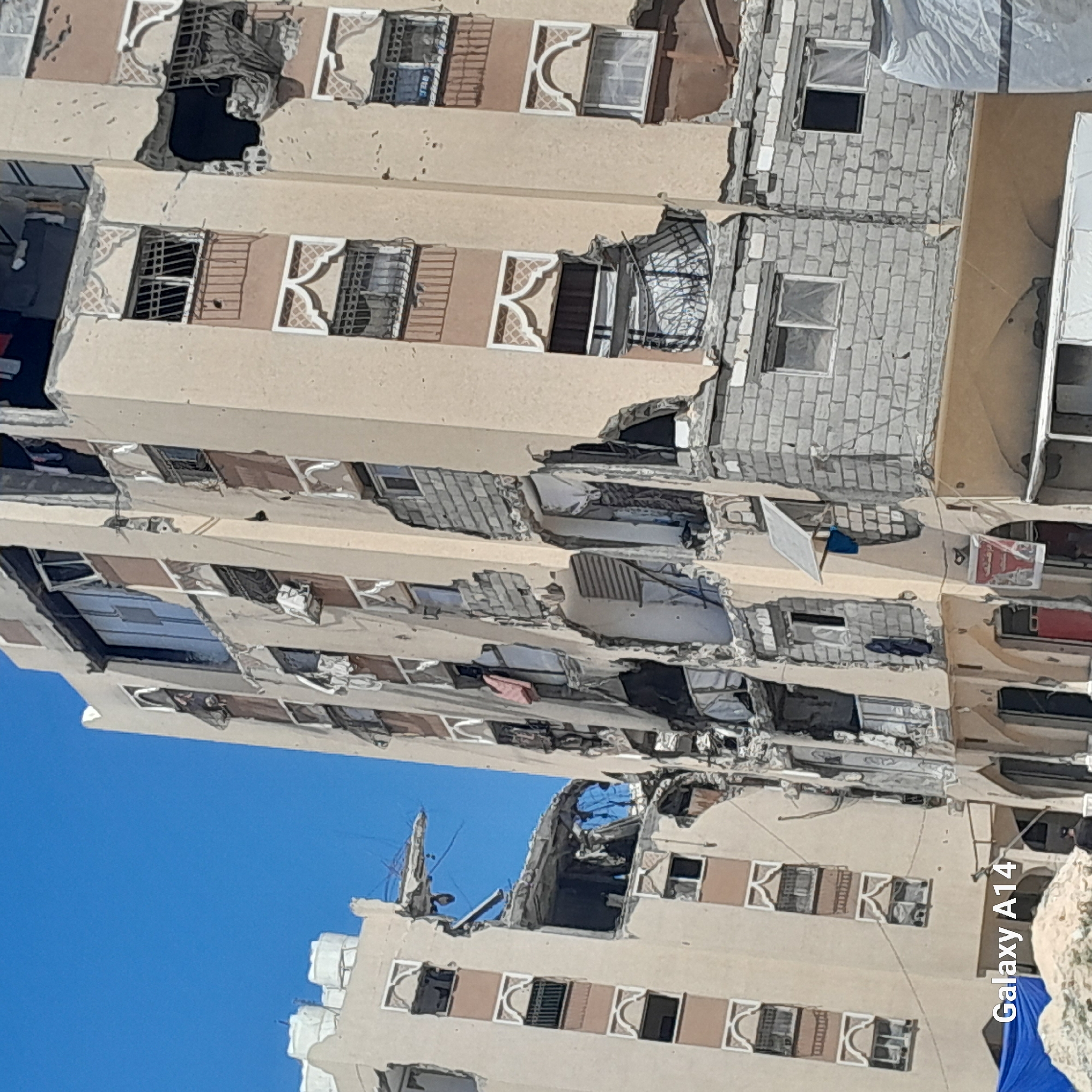 The image depicts a damaged multi-story building in Khan Yunis, Palestinian Territories, under a clear blue sky. The building exhibits significant structural damage, with large sections of its facade and interior exposed. Numerous windows are broken or boarded up, and debris is visible on several floors. Clothing hangs from some windows, suggesting recent habitation. There are also indications of external damage, such as missing sections of the building's outer walls. In the foreground, a blue tarp covers debris or rubble, and the corner of a white tarp is visible to the right. A sign with Arabic text is affixed to the lower part of the building on the right. The overall scene suggests destruction and the aftermath of an event causing severe damage to the structure.