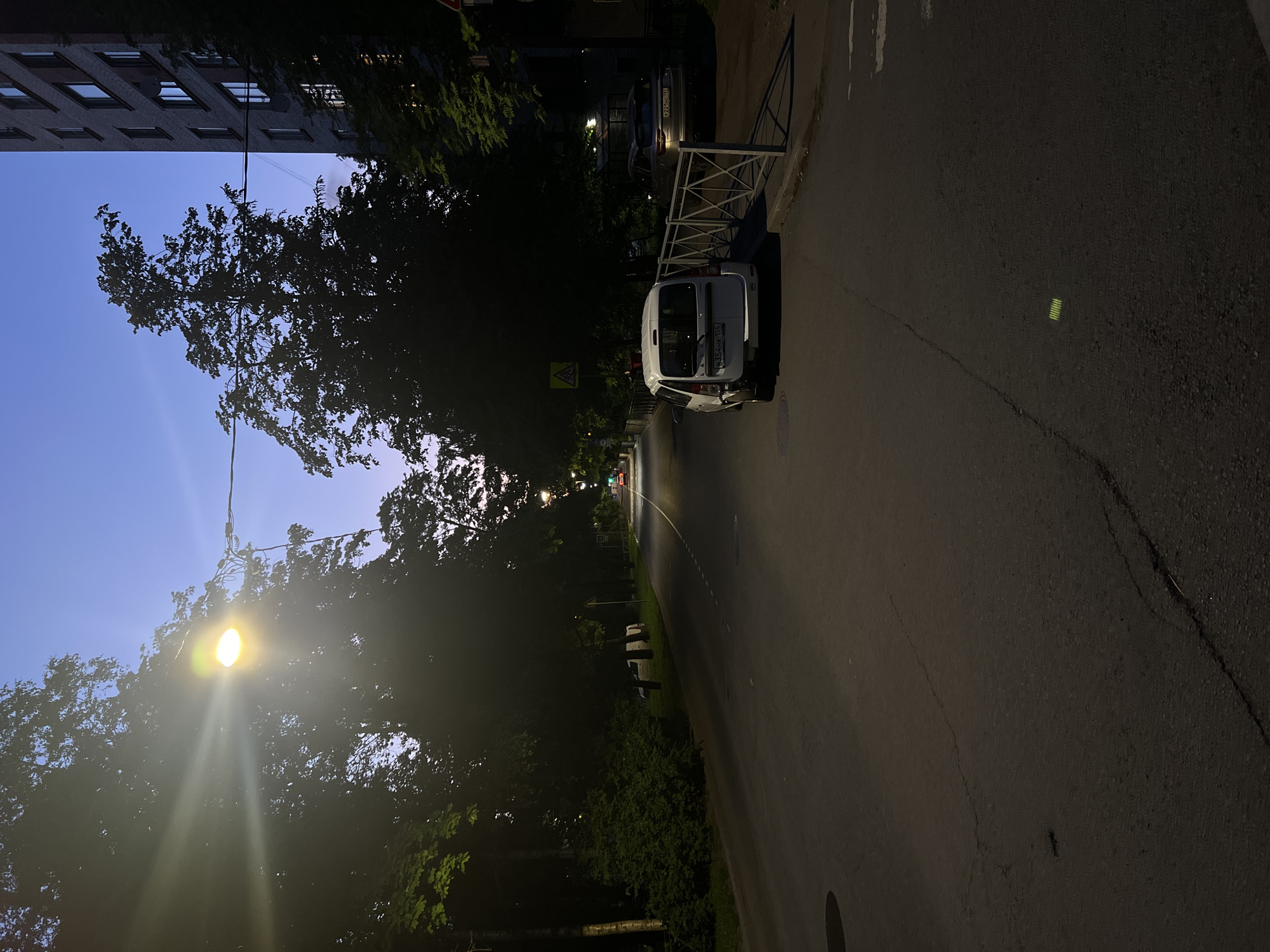 The image captures a static, dimly lit street scene at twilight in the city of Unknown, Russia. The sky overhead transitions from deep blue to a lighter, purplish hue near the horizon, indicating either dusk or dawn.
A bright streetlamp in the upper-left quadrant emits a strong, directional glare, illuminating the dark silhouette of dense trees that line both sides of the asphalt road. The road surface exhibits several cracks and features a dashed white center line extending into the distance. A manhole cover is visible on the left side of the road in the foreground, and a small, rectangular yellow reflector is present on the road surface towards the bottom-right.
On the right side of the road, a white utility van, with visible damage to its left rear bumper, is parked alongside a white metal barrier fence. The license plate, B354HK 155, indicates registration in Omsk Oblast, Russia. Directly above the van, a yellow triangular road sign displaying a children crossing symbol is affixed to a pole. Further down the road on the right, partially obscured by the fence and foliage, a silver SUV is parked. Its license plate, P221BC 31, indicates registration in Belgorod Oblast, Russia.
To the far right, a multi-story brick residential building with numerous illuminated windows is partially visible, obscured by trees. Another building structure with visible interior lights can be seen further down the road on the right. No individuals are present in the scene.