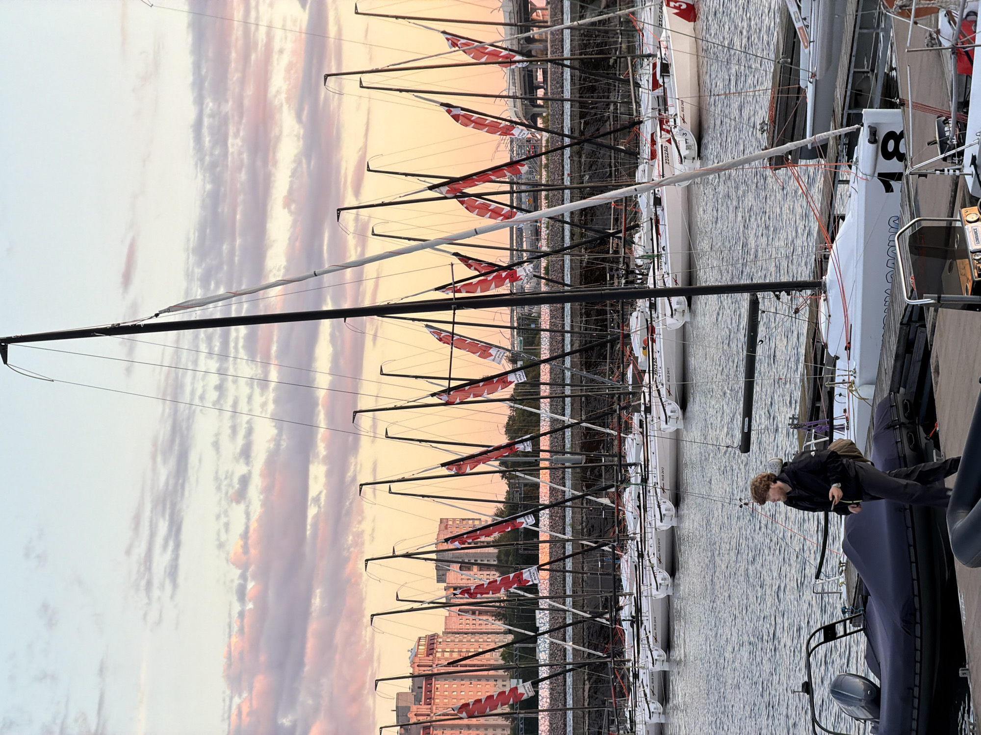In Saint Petersburg, Russia, at dusk, a person with reddish-brown hair wearing a black jacket and grey pants walks along a wooden pier next to several docked sailboats. The sky is painted with soft pink and orange hues, indicating the setting sun, and stretches above a line of moored sailboats. These sailboats, mostly white with black masts and red triangular flags bearing white markings, are arranged in rows. In the background, tall residential buildings with visible windows and balconies are illuminated by the ambient light. The scene is situated on a body of water, likely a river or canal, with a visible bridge structure in the distance.