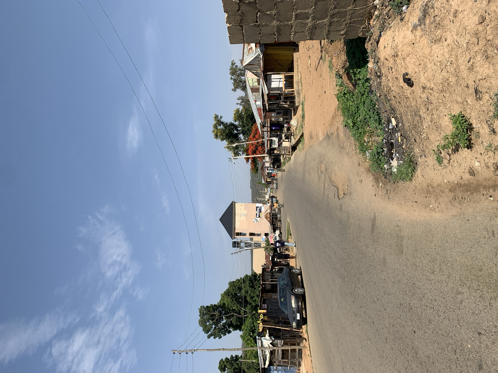 The image depicts a street scene in Rafin Par, Nigeria. The view is along a road with buildings lining both sides. On the right, there's a block structure followed by a dirt slope with litter and some greenery. The road surface is asphalt. On the left side of the road are buildings of various types, including some that appear to be shops with corrugated metal roofs. A black car is parked on the left side of the road. Several people are visible along the street near the central building. Overhead are electrical or telephone lines. The sky is blue with light cloud cover.