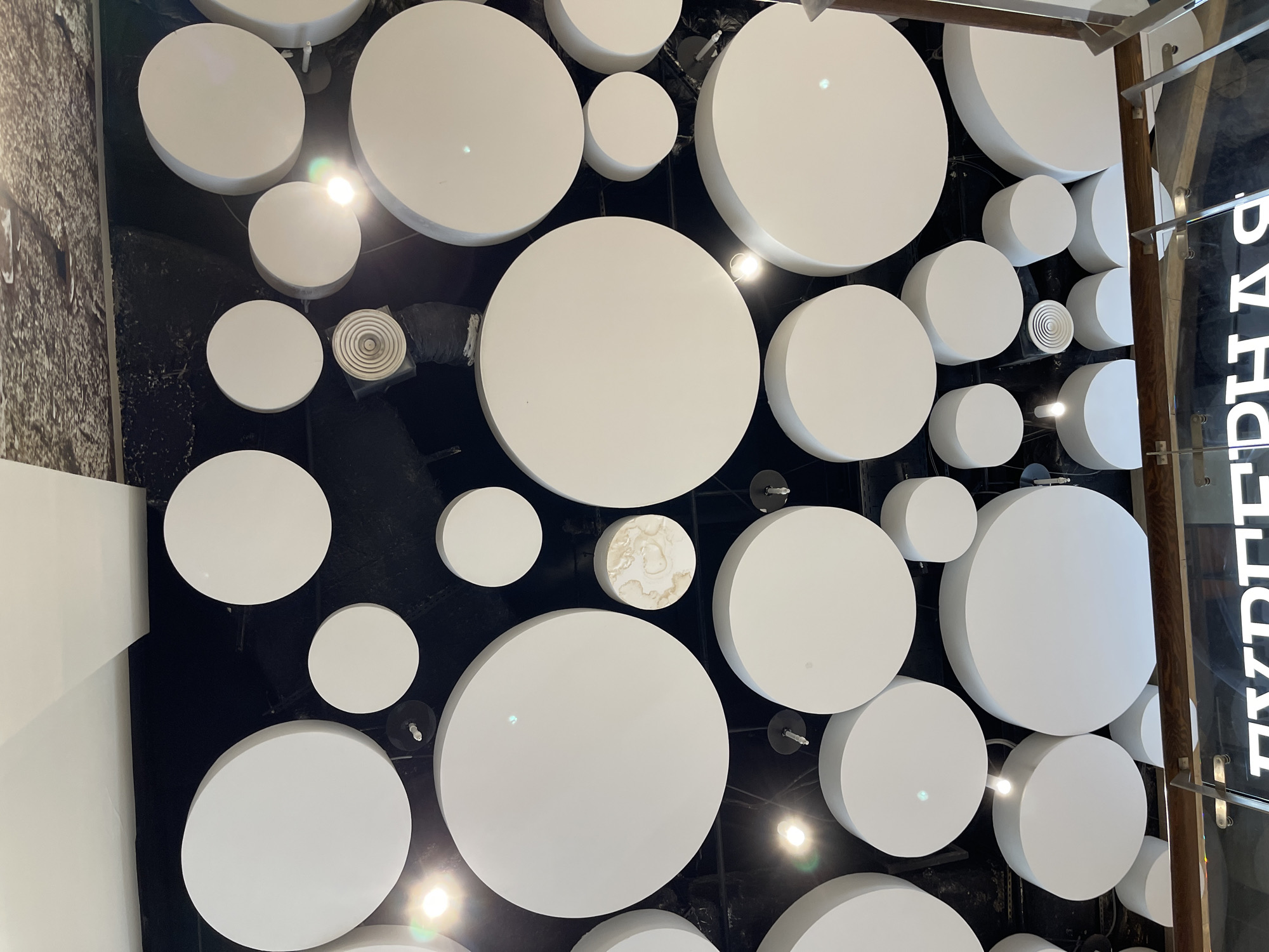 A low-angle shot of a ceiling with numerous circular white acoustic panels of varying sizes. The panels are attached to a dark background and are illuminated by spotlights, creating a play of light and shadow. Some panels have a slightly textured, marbled appearance. In the lower portion of the image, a partial view of a wooden shelf and a glass barrier with white text on it is visible. The text appears to be in Russian, possibly part of a store sign. The scene is likely indoors, in a commercial or public space, and the architectural design suggests it could be a modern shopping mall or a well-designed retail store. The image does not contain any people or visible actions.