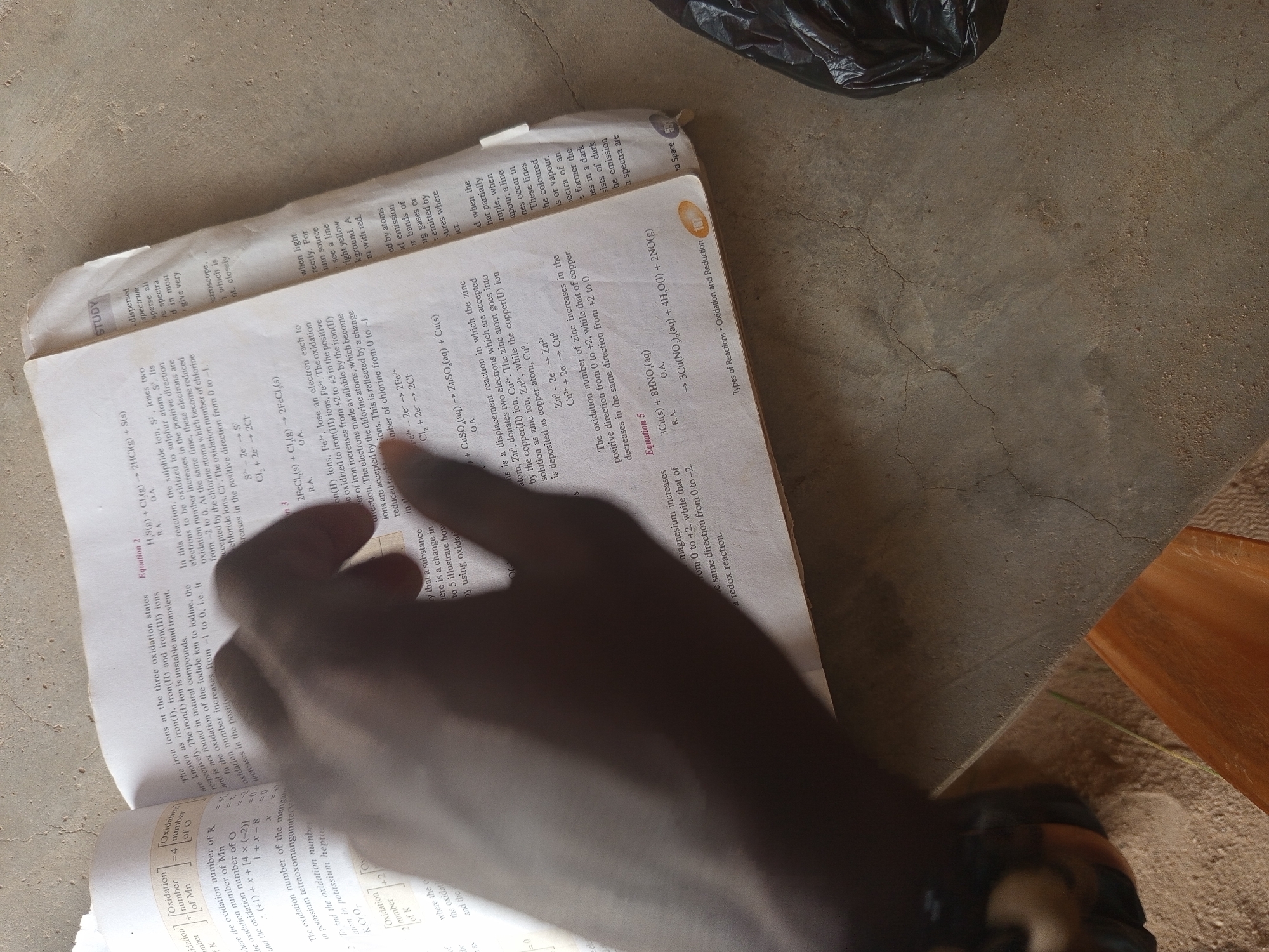 The image shows an open book lying on a textured surface, possibly concrete or a similar material. The book, which appears to be a chemistry textbook, is open to a page with chemical equations and explanatory text about oxidation and reduction. A hand is visible in the foreground, obscuring a portion of the book and the table surface. The book is positioned on a light brown, textured surface, possibly a table or floor. In the background, a dark object, possibly a bag, is partially visible. The lighting suggests an indoor setting. The overall scene is devoid of any specific city or location indicators, making it impossible to determine the city or state.
