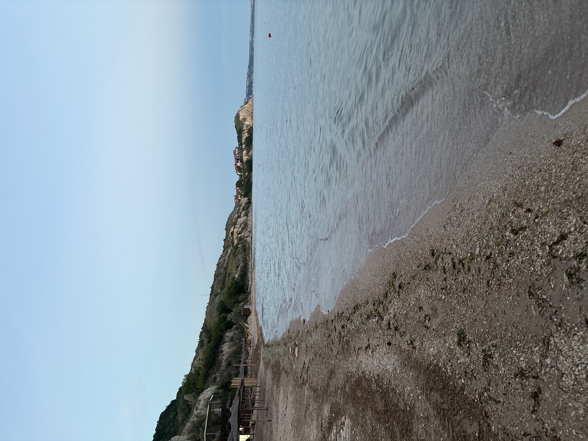 The image shows a view of a beach in Topola, Bulgaria. The beach is sandy and pebbly, with some seaweed scattered on the shore. To the left, there are several straw umbrellas and some wooden structures. The water is calm and clear, with small waves gently lapping at the shore. In the background, there is a hilly coastline with some houses visible on the clifftops. The sky is a clear, pale blue, indicating it is likely early morning or late evening. There are no people visible in the image. The overall scene is peaceful and serene, showcasing a natural coastal landscape.