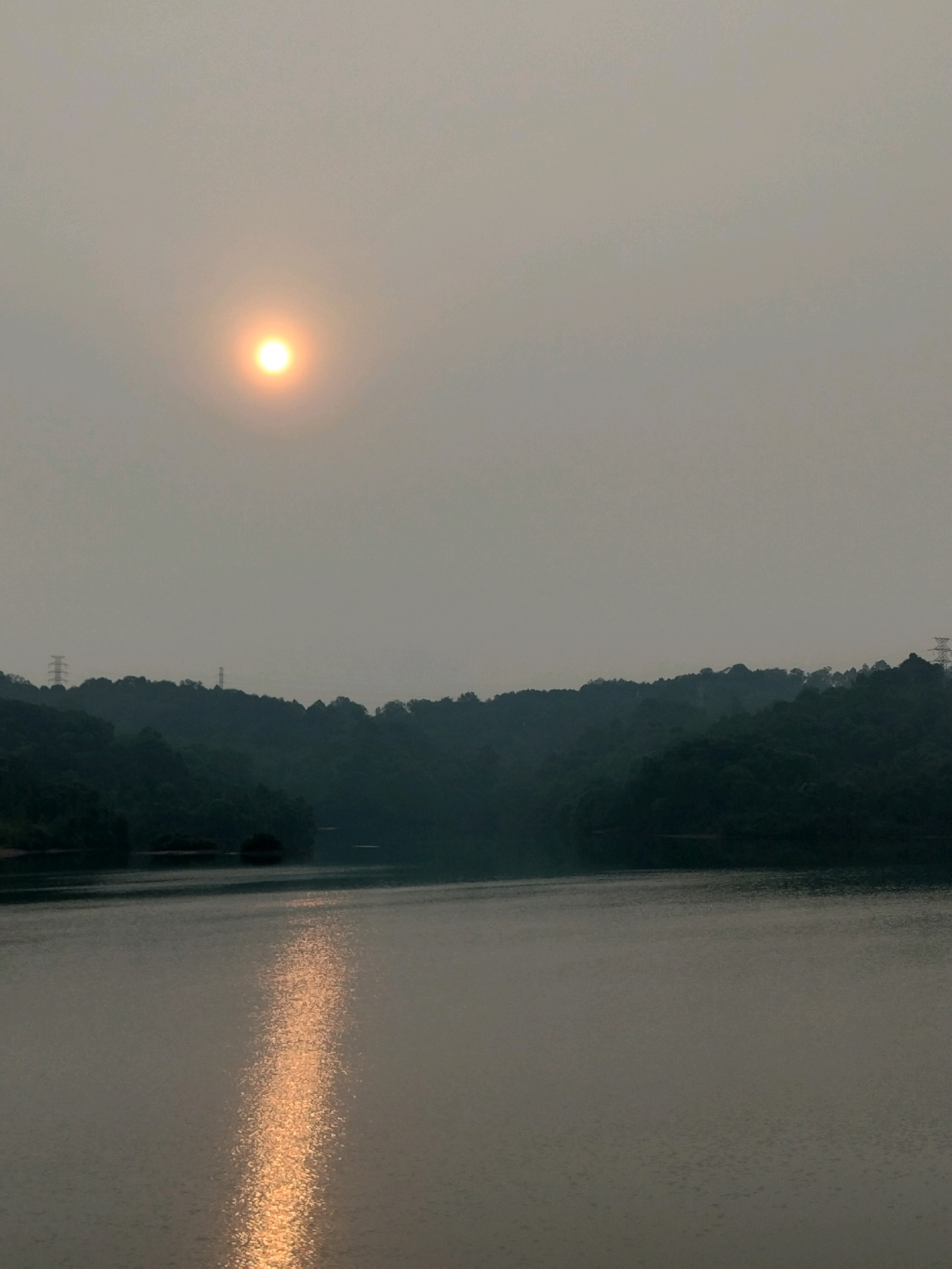 The image depicts a serene landscape, likely a reservoir or a large body of water, with a hazy sun visible in the upper left quadrant. The sun appears as a bright, diffused orb against a pale, overcast sky. In the foreground, the water surface reflects the light of the sun, creating a shimmering path of golden light that stretches towards the viewer. The middle ground is dominated by dark, forested hills that form the shoreline. The foliage is dense and appears lush, with the contours of the hills softened by the atmospheric haze. Several tall, slender electrical pylons are discernible on the crests of some of the hills, indicating the presence of human infrastructure within the natural environment. The overall mood of the image is tranquil and somewhat ethereal due to the soft light and misty atmosphere. Based on the electrical pylons and the type of forested terrain, this scene is consistent with the Nongsa district of Batam, Indonesia, possibly around the Duriangkang Reservoir. There are no people or animals visible in the frame.