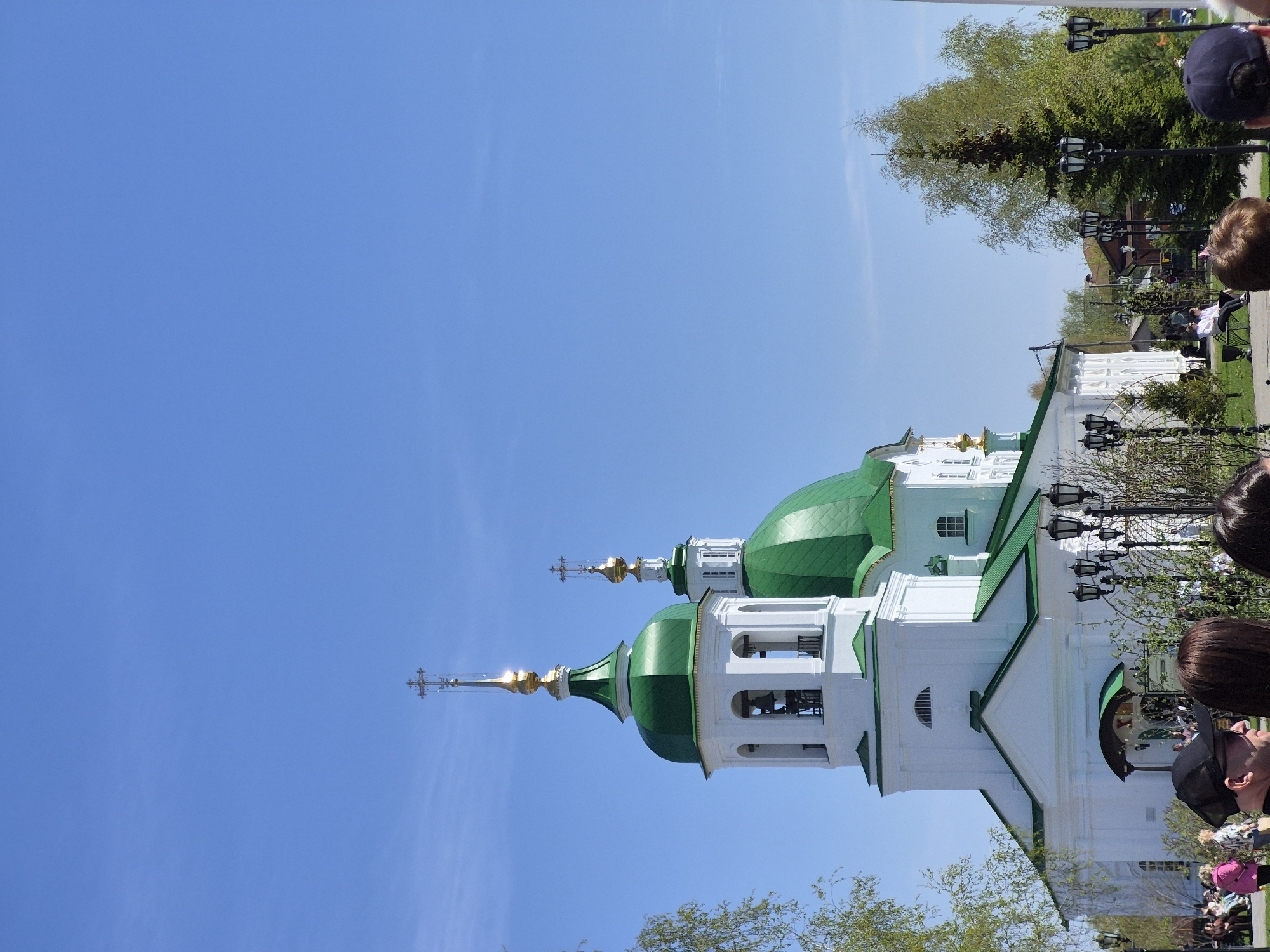 The image shows a white and green Russian Orthodox church in Tobolsk, Russia, under a bright blue sky. The church has two domes, and people are gathered in the foreground, seemingly observing the church. Street lamps and trees line the periphery of the building. A portion of a white building can be seen in the background.