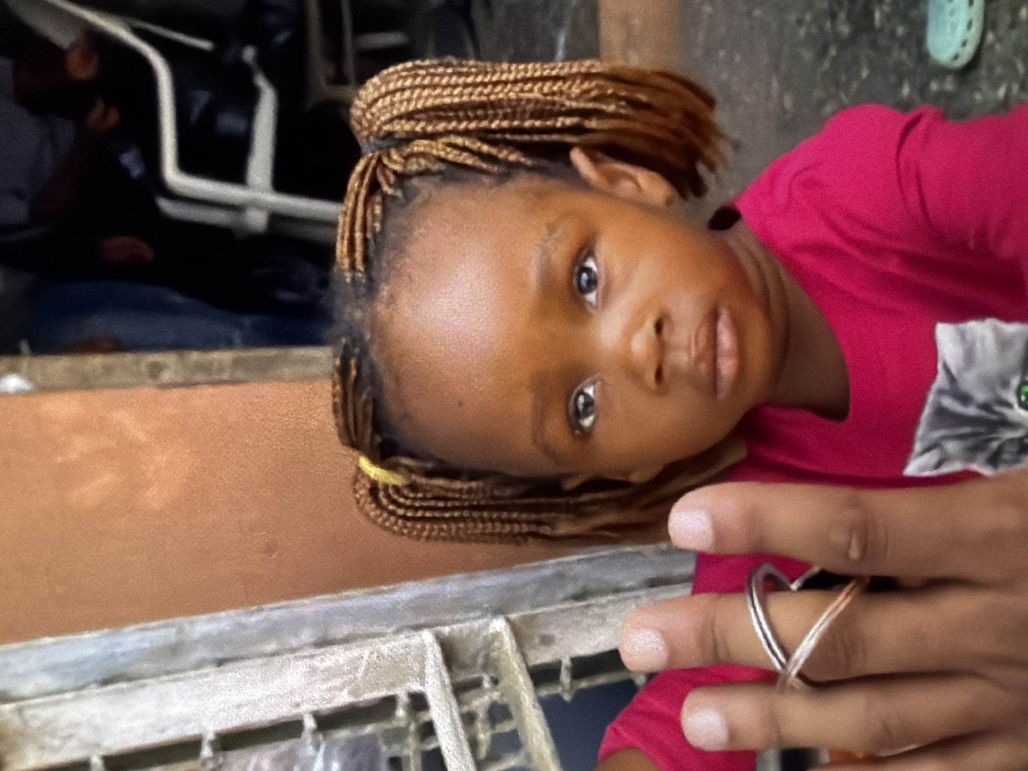 A close-up, eye-level, medium shot shows a young child of African descent with braided hair styled in two pigtails, wearing a bright pink long-sleeved shirt. The child is looking directly at the camera with wide eyes and slightly parted lips. Their left hand, adorned with a silver criss-cross ring, is held up in the foreground, partially obscuring the child's face. The background is blurred, revealing an orange-toned wall on the left and a darker area on the right with glimpses of people and furniture. The overall lighting is natural and appears to be daylight.