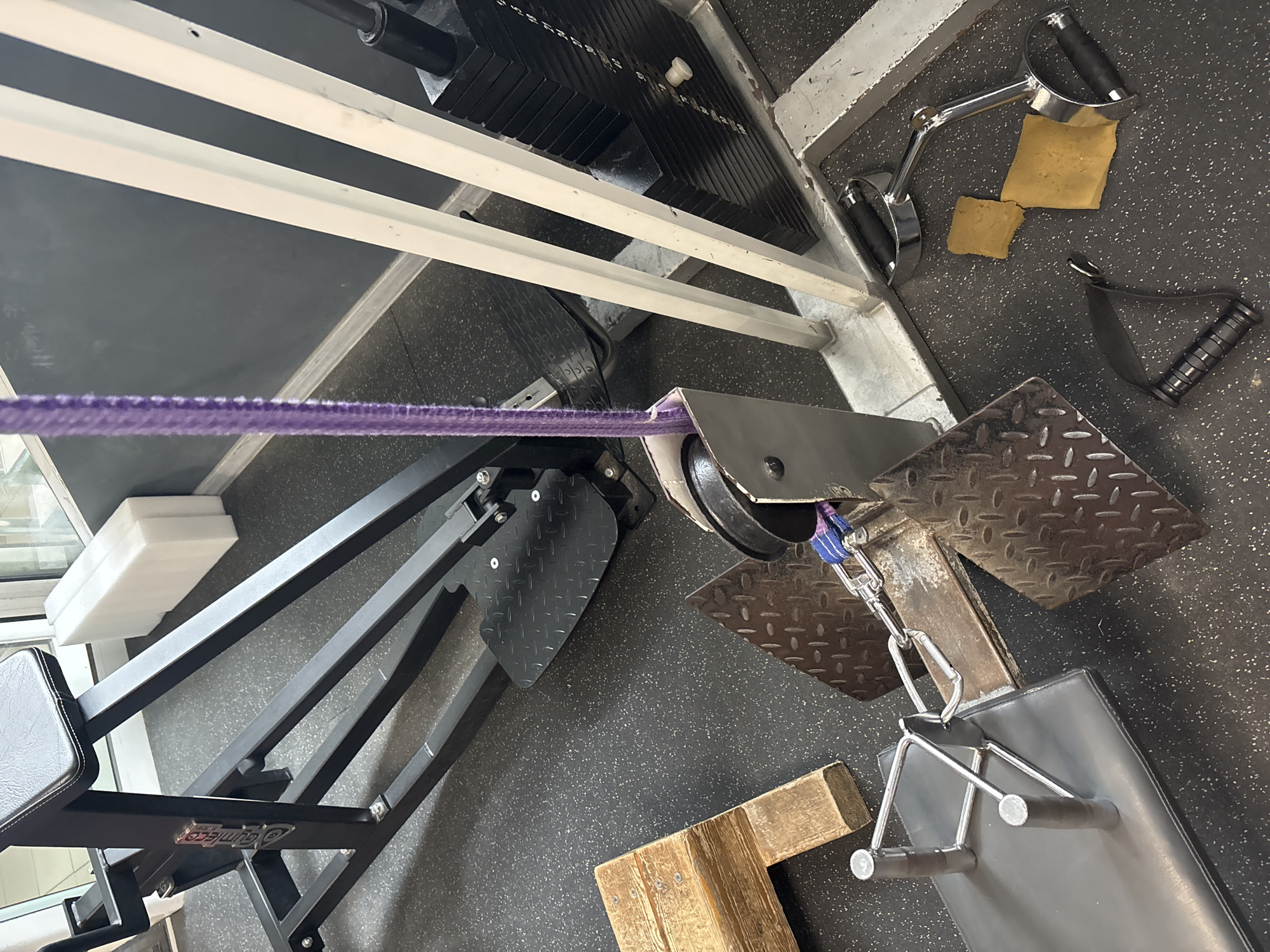 The image shows a section of a weight training machine, likely in a gym. The machine has a purple rope running through a pulley system connected to weight plates. Various handles and grips, including a V-shaped handle and single-handed grips, are seen on the floor. The floor has a black rubberized surface. There is no indication of this being in Varna, Bulgaria or involving any people.