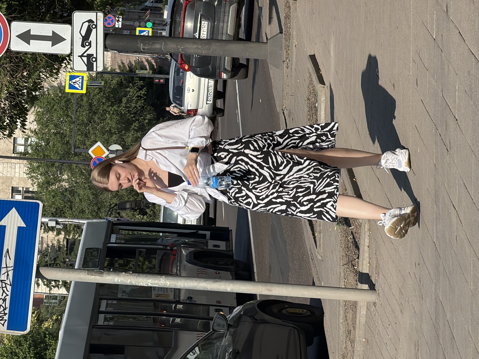 A young woman with blonde hair is walking on a sidewalk in Krasnoyarsk, Russia, on a sunny day. She is wearing a white collared shirt, a black top, and a black and white zebra-print skirt with a high slit. She has white athletic shoes and is holding a clear plastic bottle of water. She has a smartwatch on her left wrist and is wearing wireless earbuds. In the background, there is a bus, several cars, and traffic signs. The woman is walking towards the viewer.