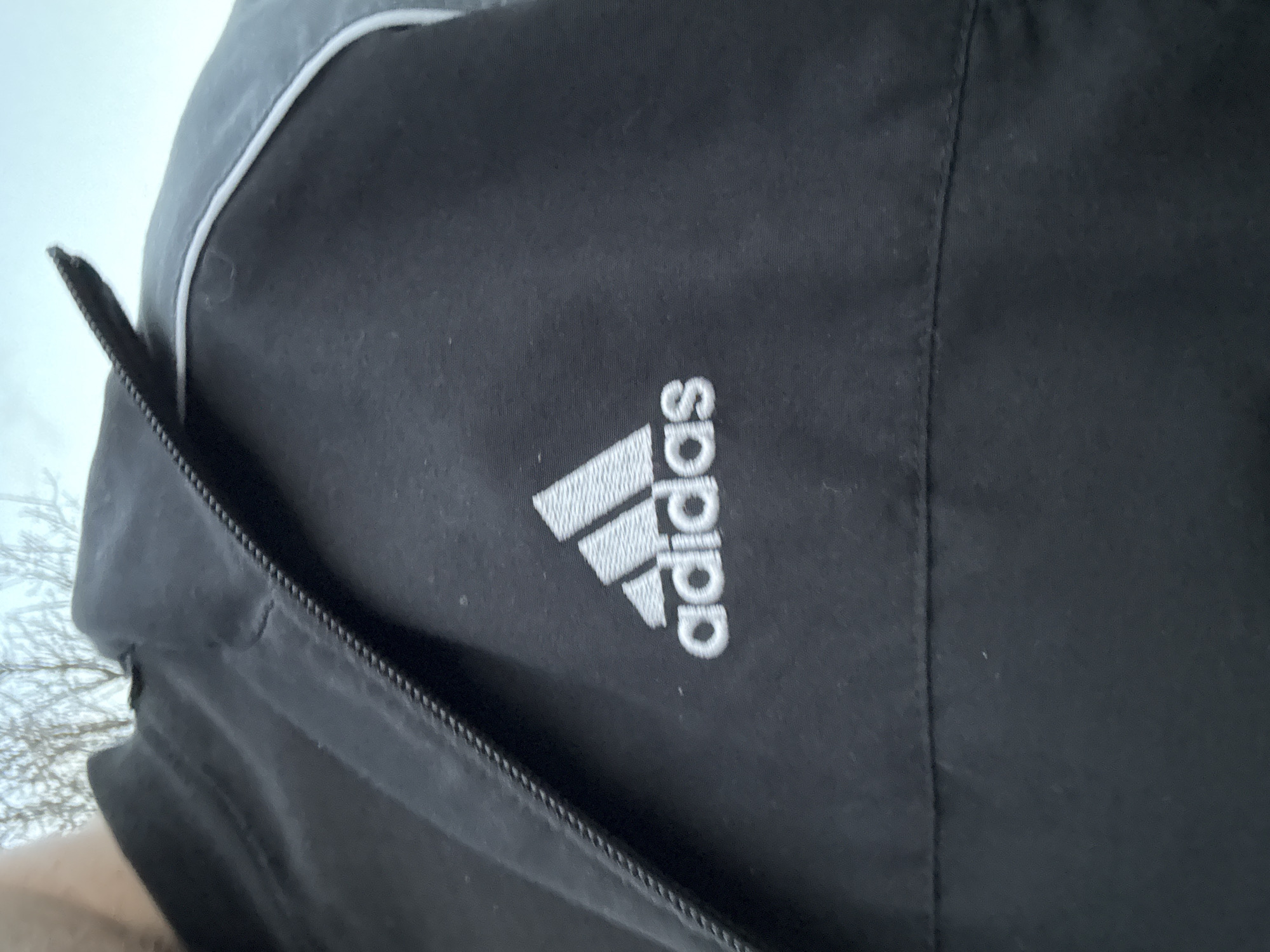 The image shows a close-up of a black Adidas jacket. The Adidas logo is prominently displayed on the jacket. A zipper is visible on the left side. The location is unknown and cannot be determined from the image.