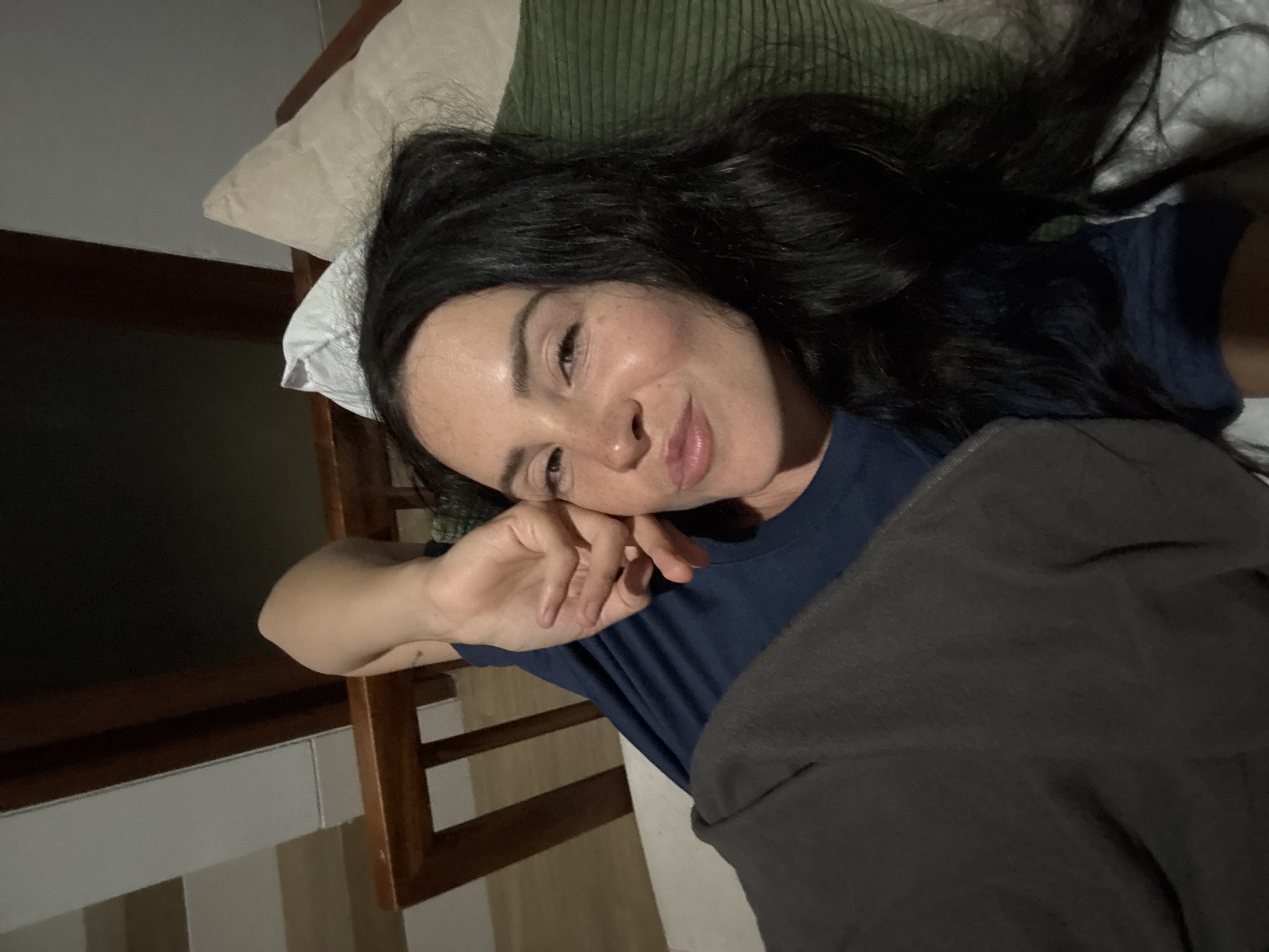 A woman with dark hair and fair skin lies on a bed, looking at the camera. She is wearing a dark blue t-shirt and is covered by a dark gray blanket. Her left hand is raised, touching her face near her eye. A green striped pillow is behind her head, and a light-colored pillow is to her right. The background appears to be an indoor setting with wooden furniture and a white wall. The lighting is dim, suggesting it is nighttime or a dimly lit room.