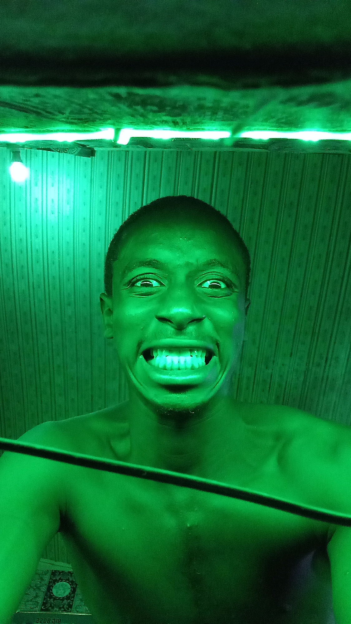 A shirtless, dark-skinned man with close-cropped hair stands indoors under green lighting, smiling broadly and showing his teeth. He is looking upward at the camera. Behind him is a striped wall, with a light fixture and a strip of green lighting on the ceiling. A dark cord stretches across the lower portion of the image. The location is stated to be in Onitsha, Nigeria.
