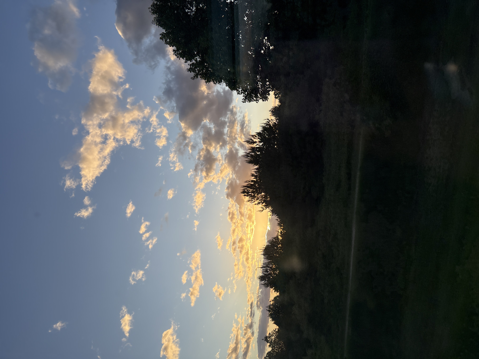 The image shows a sunset in Мажэйкаўскі сельскі Савет, Belarus. The sky is a mix of blue and orange, with scattered clouds that are lit by the setting sun. Trees are visible along the horizon line. The landscape seems to be dark and green. There are no people visible.