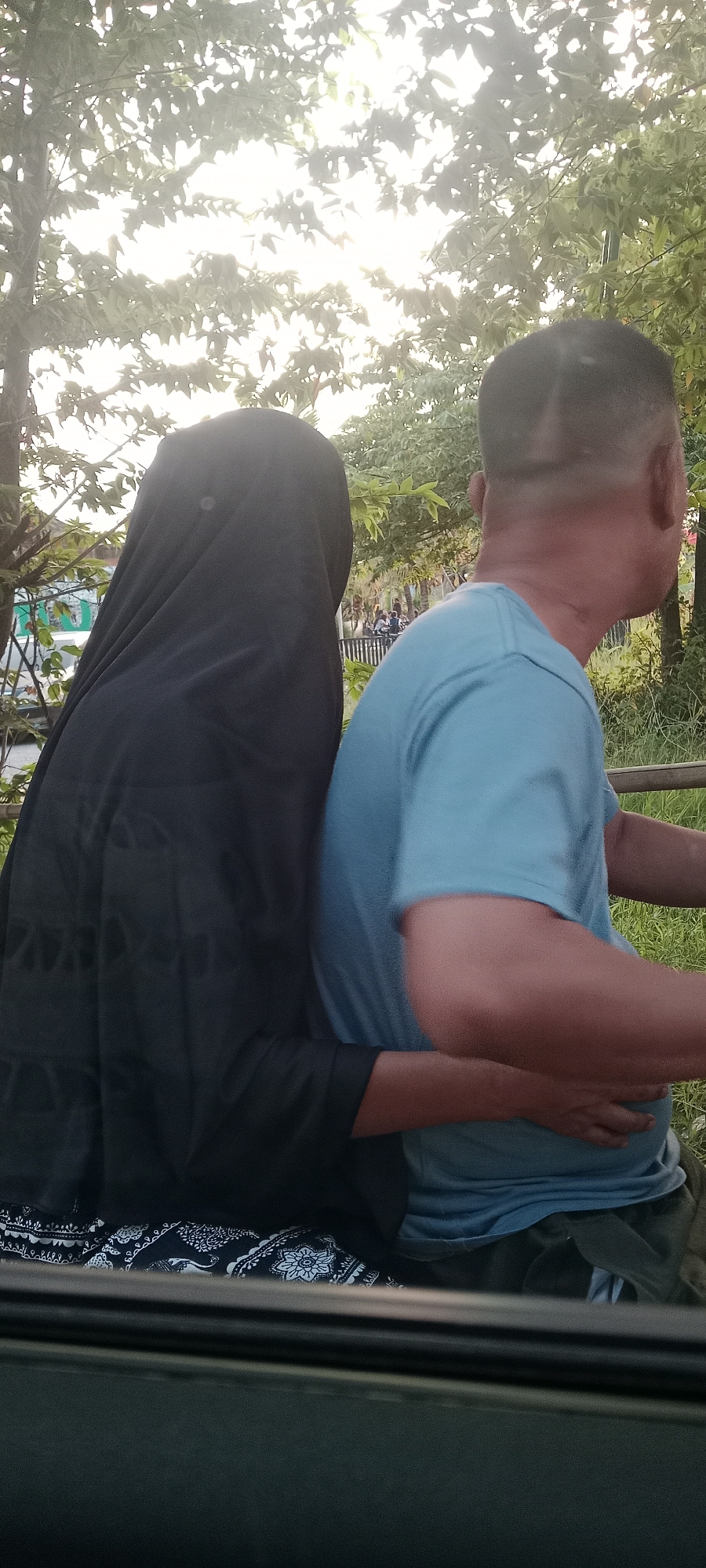 Two individuals are seated side-by-side in what appears to be an outdoor setting, possibly near a park or a roadside in Banyuraden, Indonesia, as suggested by the foliage and lighting. The person on the left is fully covered in a black hijab, obscuring their face and most of their body, with only their hands visible. They are wearing patterned pants with a black and white design. The person on the right, identified as male by their haircut, wears a light blue t-shirt and dark green pants. Their right arm is wrapped around the waist of the individual in the hijab, and their left hand rests on their own lap. The background shows dense green foliage and trees with sunlight filtering through, indicating daytime. A railing or fence is visible horizontally across the middle of the image.