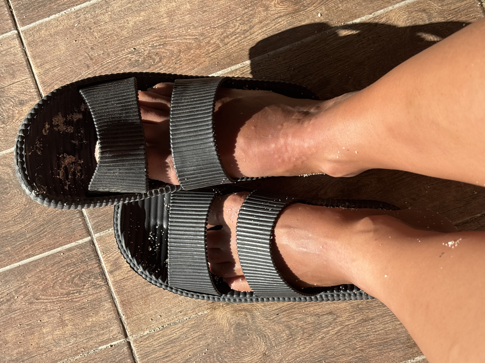 A high-angle shot shows a pair of feet in black rubber sandals resting on brown, wood-patterned tile flooring. Fine grains of sand are visible on the skin and sandals. The location is Kuta Selatan, Indonesia.