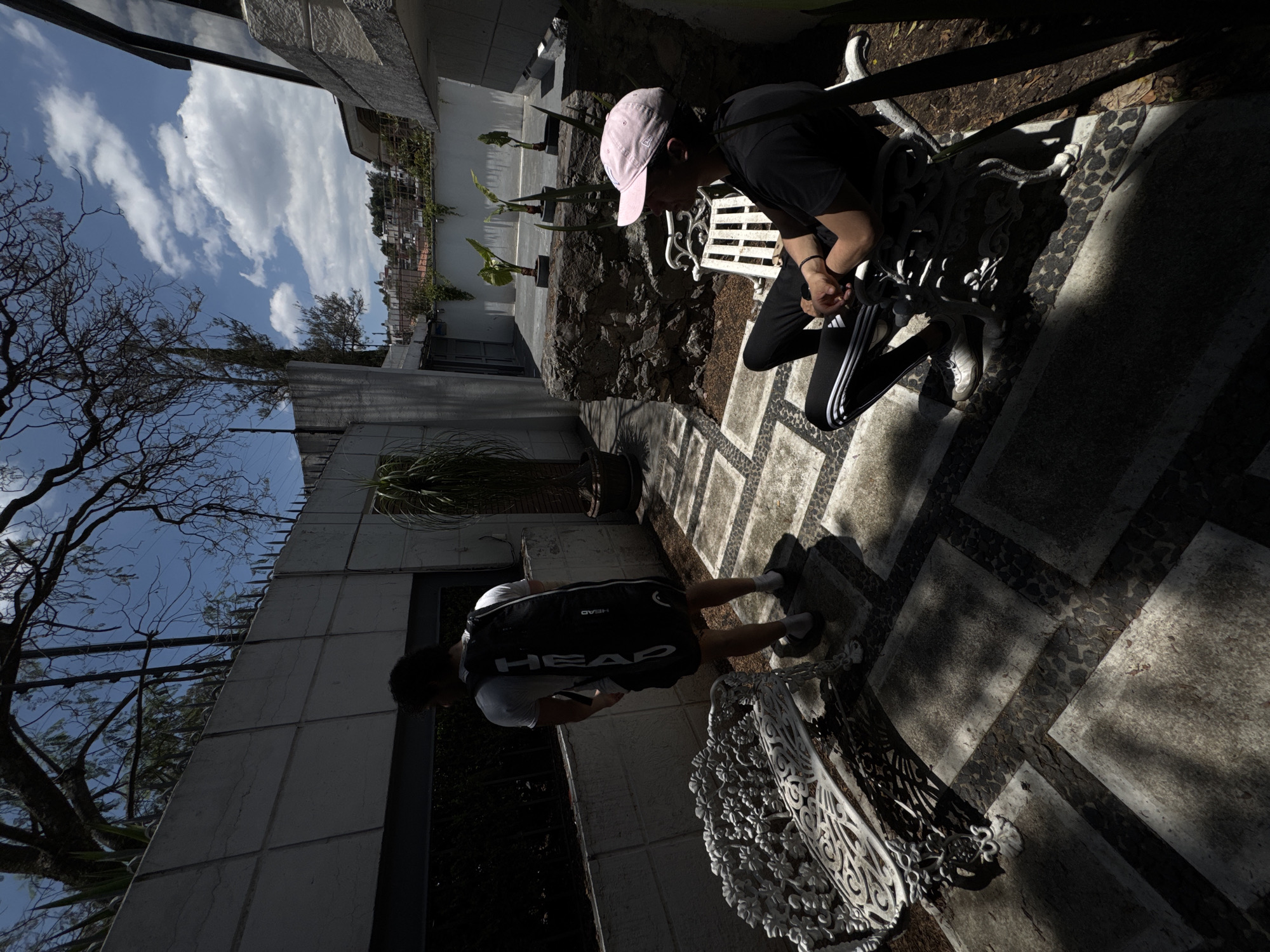 This image depicts an outdoor courtyard or patio area under bright daylight, featuring two individuals, architectural elements, and vegetation. The strong, directional sunlight creates prominent shadows across the scene.
In the right foreground, a person wearing a pink baseball cap, a black t-shirt, and black Adidas track pants with distinctive white stripes is seen squatting or bending over. Their hands are clasped together, suggesting they are either adjusting something or resting. They wear white sneakers, and next to them is a white, ornately designed metal bench.
Further back on the left, another individual stands with their back largely to the viewer. They are dressed in a white t-shirt and black shorts, carrying a black backpack emblazoned with the word "HEAD" in white capital letters. A portion of a similar white ornamental bench is visible near their feet.
The ground is paved with a geometric pattern of light and dark textured squares and rectangles, over which the sharp shadows extend. Along a low wall in the mid-ground, several tall, potted plants with slender leaves are arranged. In the background, a large tree with intricate, bare branches stands against a clear blue sky, dotted with fluffy white clouds. Parts of buildings and a distant urban landscape can be seen. The architectural style and presence of a bare-branched tree (potentially a jacaranda common to Mexico City) align with the provided location context. The overall atmosphere is casual and relaxed.
Visible text includes the brand "HEAD" on the backpack and the implied "ADIDAS" from the characteristic stripes on the pants.