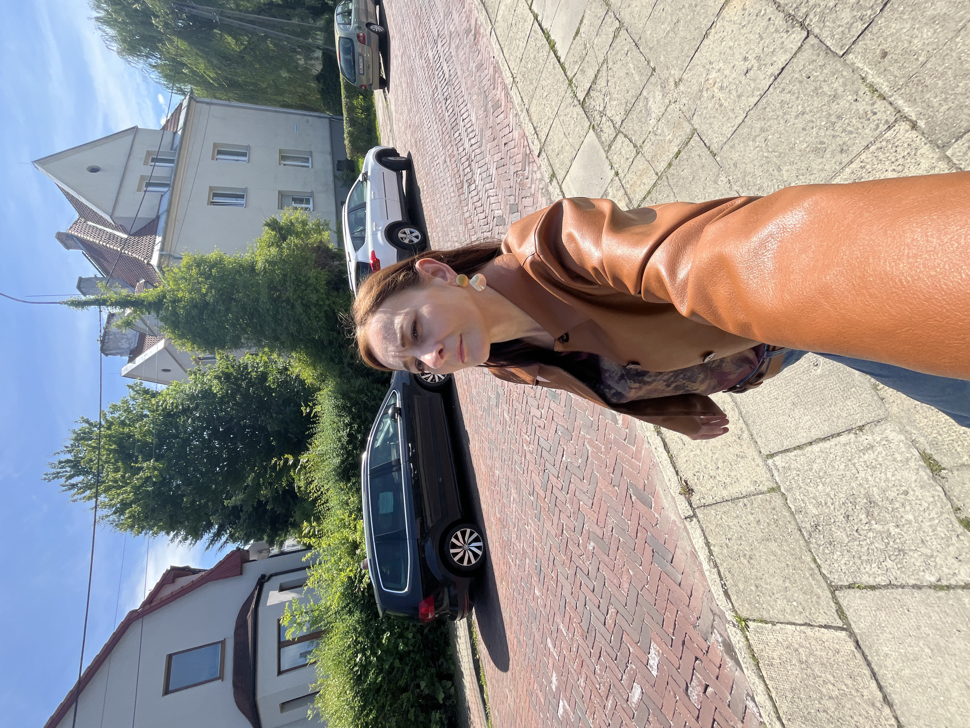 A woman with auburn hair is taking a selfie on a sunny day on a street in the city of Unknown, Unknown. She is wearing a brown leather jacket over a patterned shirt and blue jeans. She has large, round earrings. The street is paved with red bricks and lined with stone sidewalks. Behind her, several cars are parked on the street, including a black minivan, a white SUV, and a light-colored sedan. Residential buildings with red-tiled roofs and trees are visible in the background. The sky is blue with scattered clouds.