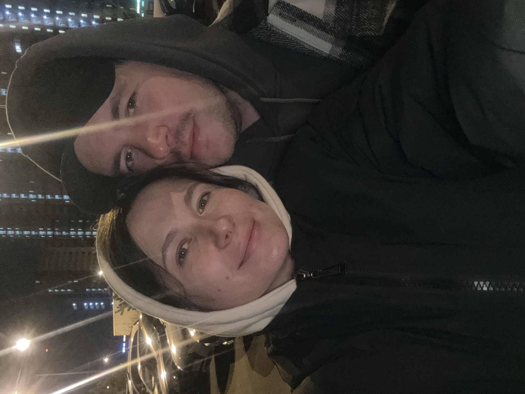 A nighttime selfie captures two individuals, a female and a male, in a close-up shot within an urban environment in the city of Odintsovo, Russia. The female, positioned on the left, has light skin, dark hair, and exhibits a slight smile, looking directly at the camera. She wears a light-colored hooded sweatshirt with the hood up, covered by a dark, possibly black, zippered jacket. The male, positioned behind and to the right of the female, also has light skin and facial hair (stubble). He wears a dark hooded sweatshirt with the hood up, layered with a plaid jacket featuring grey, white, and black patterns, and is also looking towards the camera. The background consists of a parking area with multiple dark-colored vehicles, dimly lit by artificial light. Distant, multi-story residential buildings with illuminated windows are visible. Several streetlights provide illumination, with one prominent light source in the upper left of the frame creating a lens flare.
