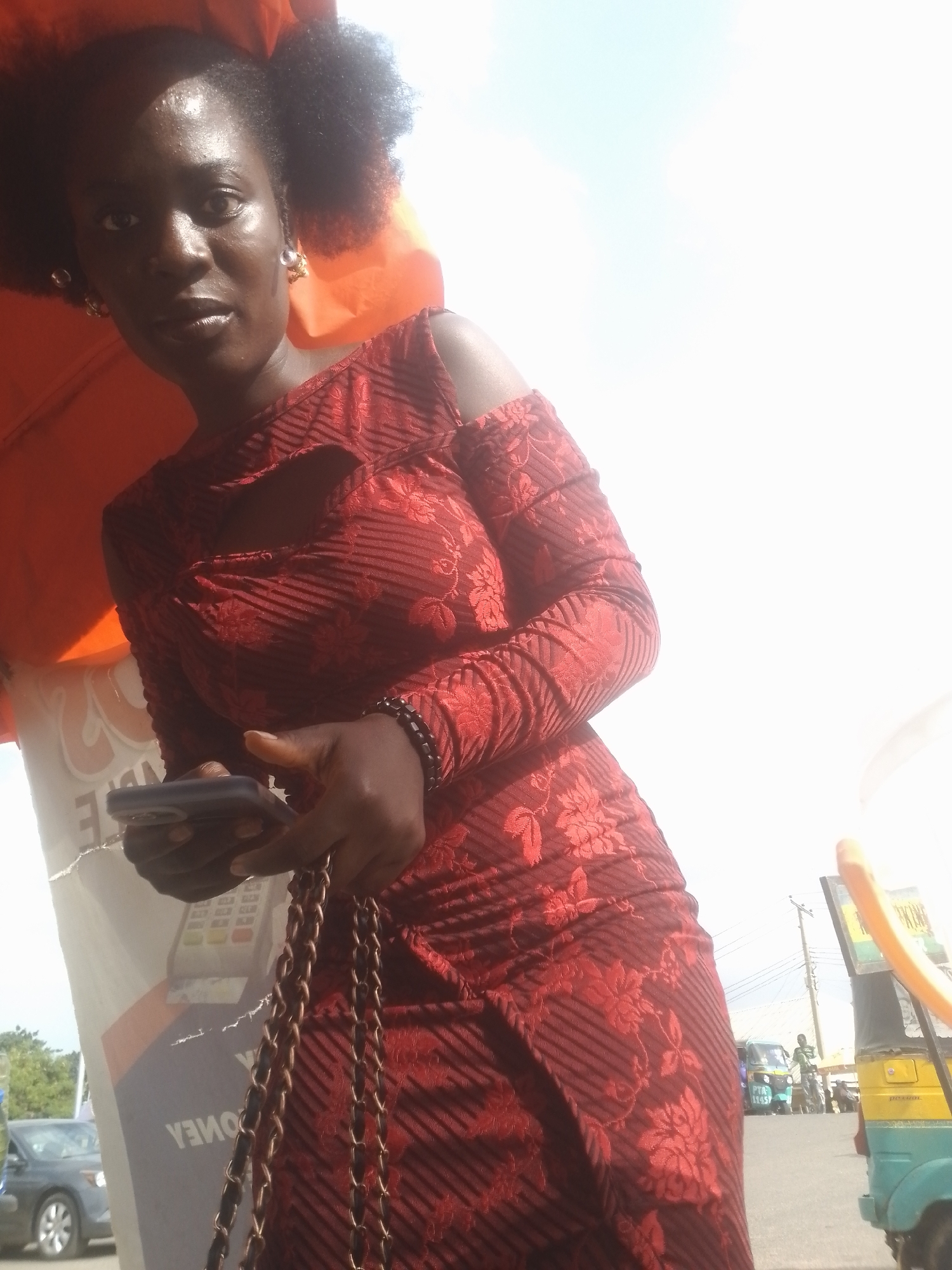 A medium shot captures a woman in a red dress adorned with floral patterns and subtle stripes. She holds a dark-colored smartphone in her left hand, while her right hand rests on the phone. A gold-colored chain strap of a bag is draped over her right shoulder. Her dark, curly hair is styled into two puffs, and she wears large, pearl-like earrings. The woman's skin appears slightly oily, reflecting the bright sunlight. In the background, an orange awning partially covers the top left of the frame. A sign with text, partially obscured and featuring a credit card reader, is visible behind her left shoulder. The scene appears to be outdoors, with a street and several vehicles, including a yellow and blue auto-rickshaw, in the distance. The sky is bright and overcast.