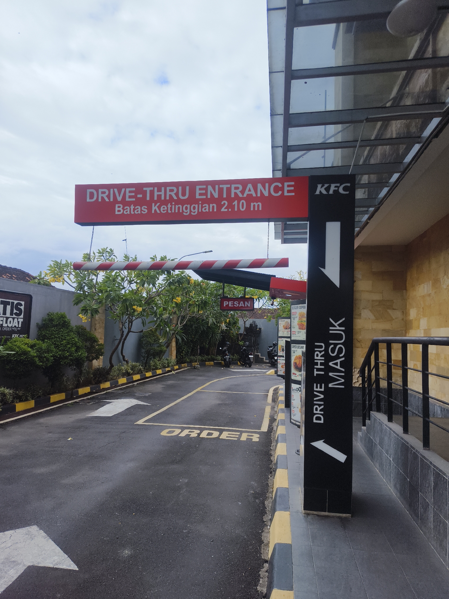 This is a photograph of a KFC drive-thru entrance in Denpasar, Indonesia. The image is taken from a low angle, looking up at a red sign that reads "DRIVE-THRU ENTRANCE Batas Ketinggian 2.10 m". Below the sign is a black pole with a white arrow pointing downwards and the words "DRIVE THRU MASUK" written vertically. The drive-thru lane is paved with asphalt and marked with yellow lines and arrows indicating the direction of traffic. There are trees and bushes along the side of the lane, and a building with a beige stone facade in the background. The sky is overcast with some clouds. In the foreground, there is a yellow and black striped barrier at the entrance to the drive-thru. A sign on the left side of the image reads "TIS FLOAT". Several motorcycles are parked to the left of the drive-thru lane, and a menu board with food items is visible on the right.