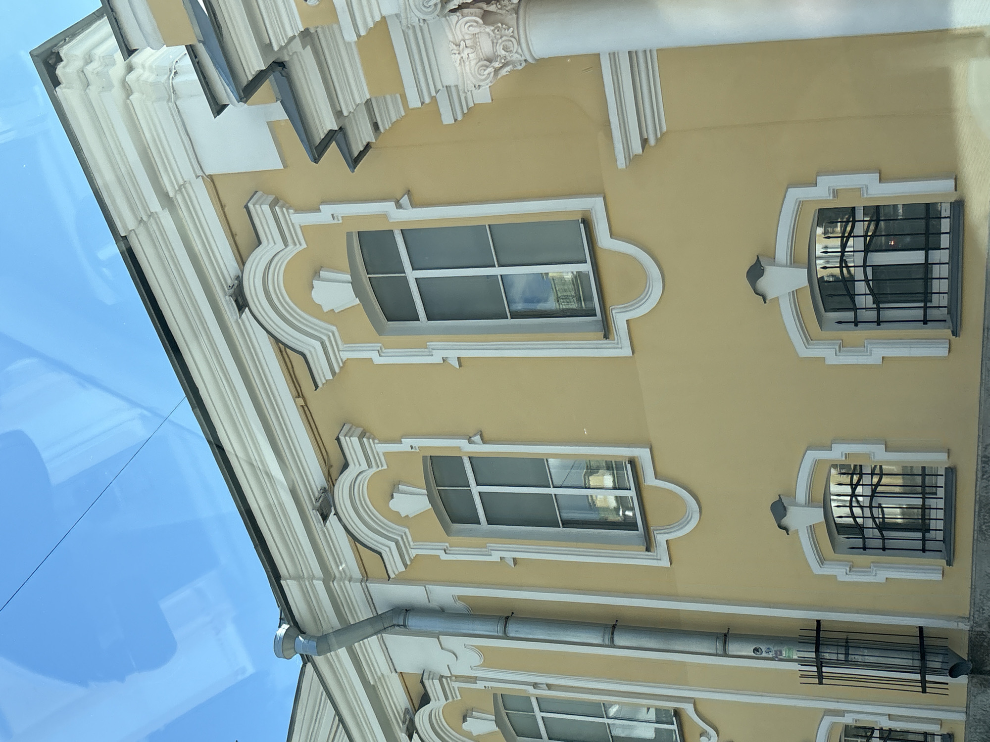 The image shows a section of a pale yellow building with white architectural details. The building has several windows, some with decorative white surrounds and metal grates. A downspout runs vertically along the left side of the building. The sky above is blue with some reflections visible on what appears to be a glass surface, possibly a window from which the photo was taken. The architectural style suggests it could be located in Saint Petersburg, Russia, a city known for its historic buildings with similar embellishments. The scene appears to be an exterior view of the building.