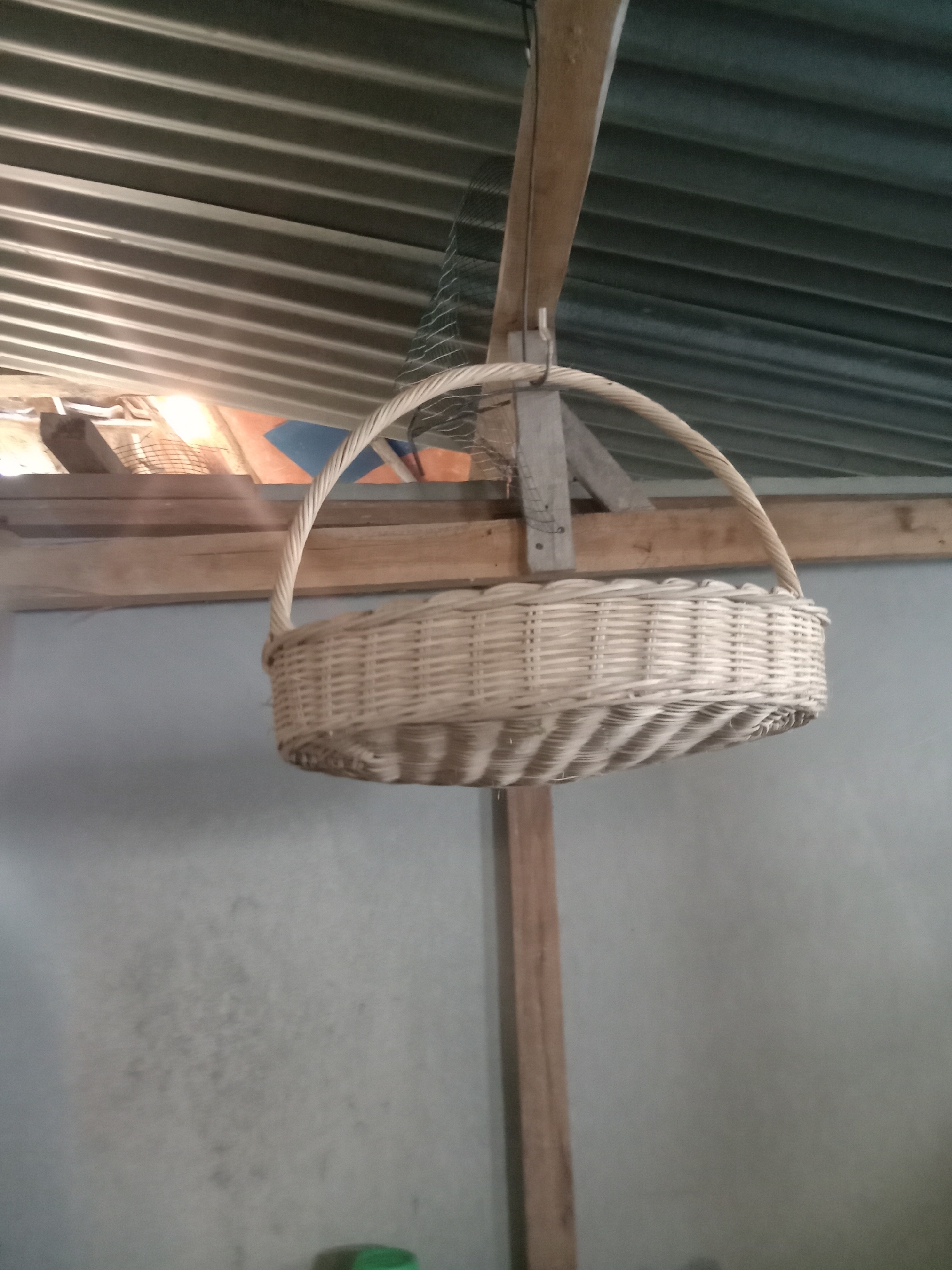 The image shows a woven basket suspended from a wooden beam under a corrugated metal roof. The basket is empty and appears to be made of light-colored wicker. It has a curved handle. Wooden planks are visible as part of the structure supporting the roof, and a light gray concrete wall is in the background. The scene is likely located in a rural or semi-rural setting within Stiaji, Indonesia, possibly a barn, storage area, or an outdoor shelter. There are no people or animals present in the image.