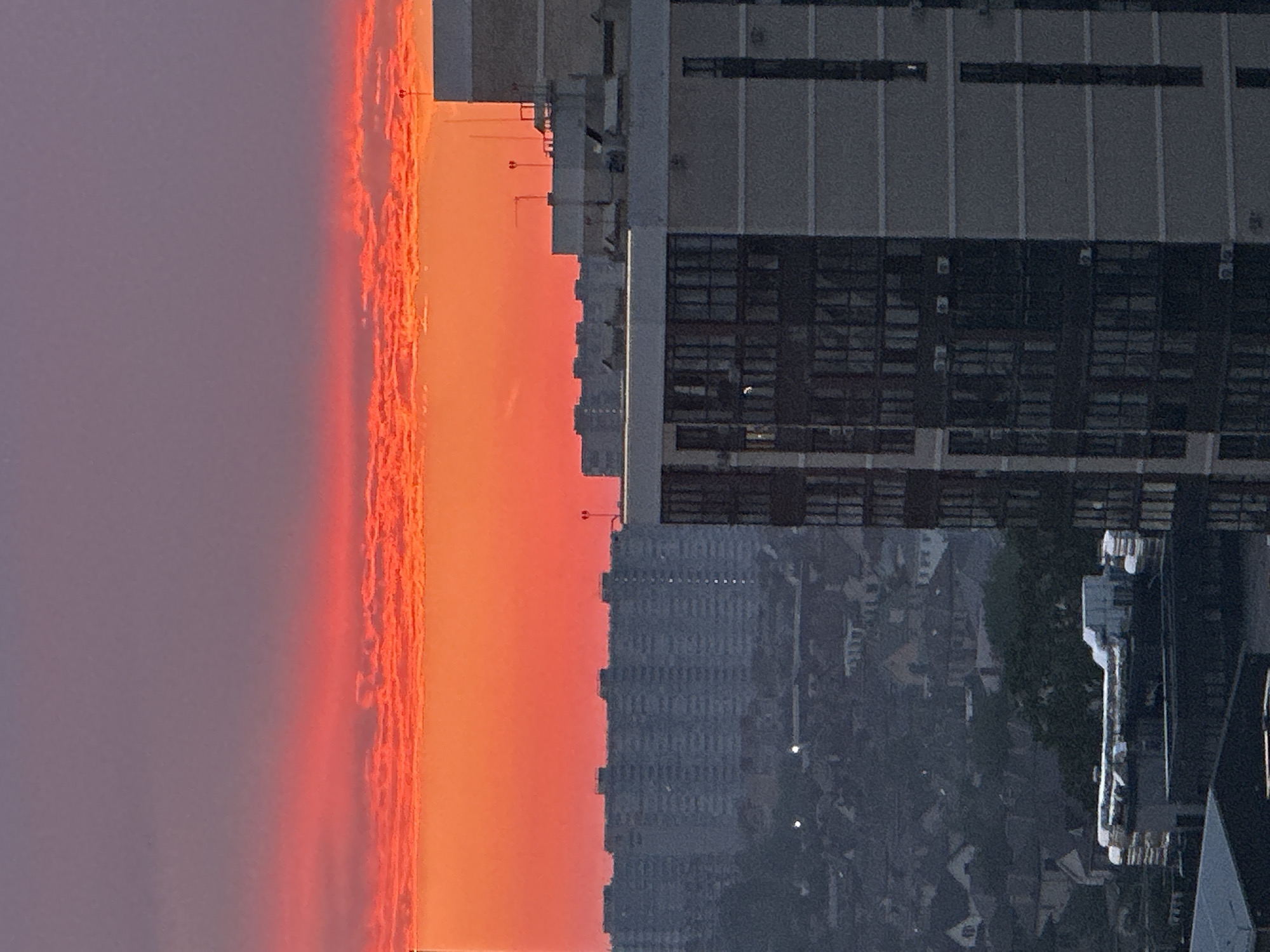 The image captures a vibrant sunset over a cityscape. The sky is dominated by fiery orange and red hues, with streaks of pink and purple clouds. Below the sky, a line of tall, grey apartment buildings stretches across the horizon, their numerous windows reflecting the fading light. In the foreground, a modern, multi-story building with dark windows and light grey exterior panels is prominently featured. The lower portion of the image reveals a cluster of smaller, less distinct buildings and trees, suggesting a mix of residential and urban development. The overall impression is one of a dynamic and colorful end to the day in a densely populated urban environment. The image does not contain any discernible people or specific actions. Based on the architectural style of the apartment buildings, which are characteristic of Soviet-era mass housing, and the general appearance of the cityscape, this scene could be representative of many urban areas in Russia, including Krasnodar.