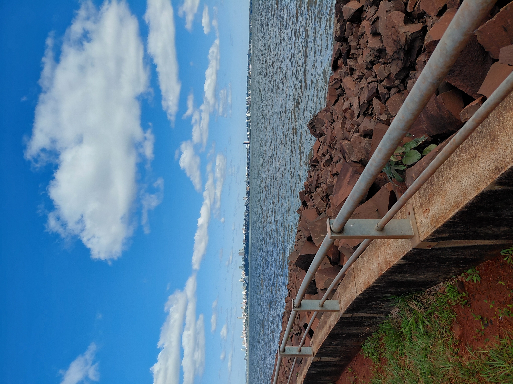 The image captures a waterfront scene in Encarnación, Paraguay. In the foreground, a concrete retaining wall lines the edge of the land, topped by a metal railing. To the right of the wall, a pile of large, reddish-brown rocks forms the embankment leading down to the water. Sparse green grass grows on the dirt to the left of the wall. The water, appearing choppy and dark blue, stretches towards a distant cityscape. The skyline of Encarnación is visible across the water, characterized by numerous buildings of varying heights, some appearing as white towers. The sky above is a vibrant blue, dotted with scattered white cumulus clouds. The overall impression is a scenic view of the city's waterfront on a clear day.