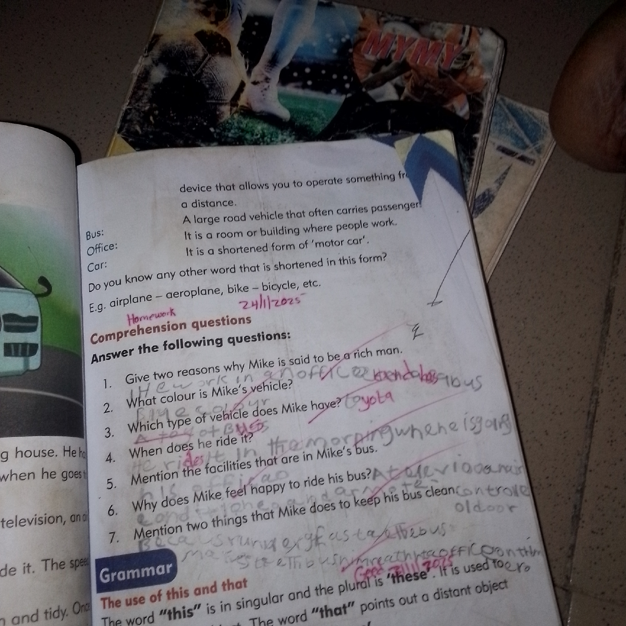 The image displays an open book with handwritten notes and markings. The book appears to be a children's textbook, featuring printed text and possibly illustrations, one of which is partially visible on the left showing a blue car. The text on the right-hand page defines various modes of transportation like "Bus," "Office," and "Car." It also presents "Comprehension questions" with numbered items and handwritten answers. For example, question 1 is "Give two reasons why Mike is said to be a rich man," and question 2 asks "What colour is Mike's vehicle?" Handwritten answers are scrawled next to these questions. A date, "24/11/2025," is written on the page, as is a heading "Grammar" with explanations about the usage of "this" and "that." The overall scene is a close-up of the book on a textured surface, possibly a table or floor. There is no discernible information within the image that pertains to the city of Jalingo, Nigeria.
