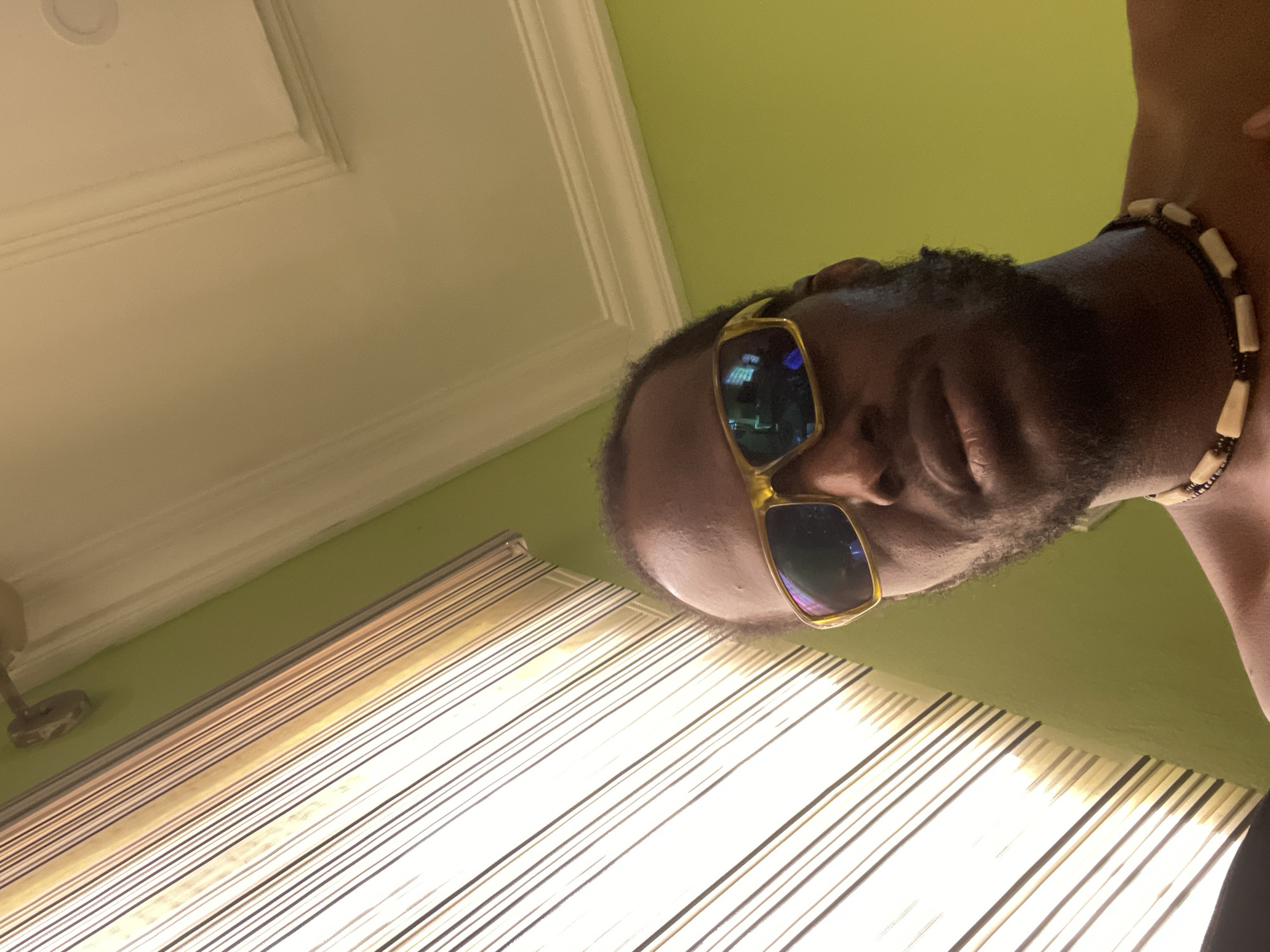 A close-up, tilted shot focuses on a dark-skinned man wearing gold-framed sunglasses and a beaded necklace. The background features a striped window covering, a beige ceiling with molding, and a green wall. The location is most likely indoors. There is no evidence to suggest the location of the city of Abuja, Nigeria.