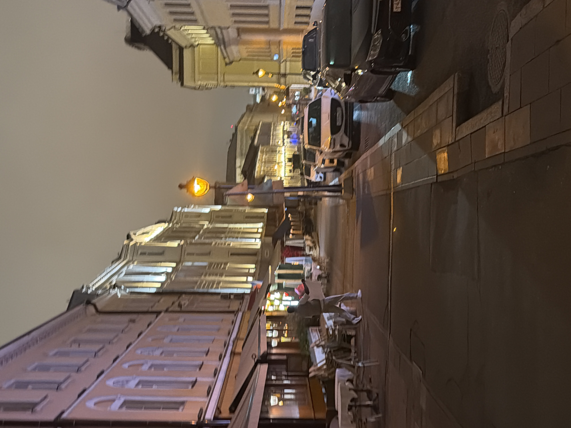 A nighttime street scene in Moscow, Russia, shows a street lined with buildings, some lit up. Vehicles are parked along the right side of the road. A man wearing light colored clothing and a pink hat stands near tables and chairs set up on the sidewalk, appearing to be stacking or arranging them. The sky is overcast, and streetlights illuminate the area.