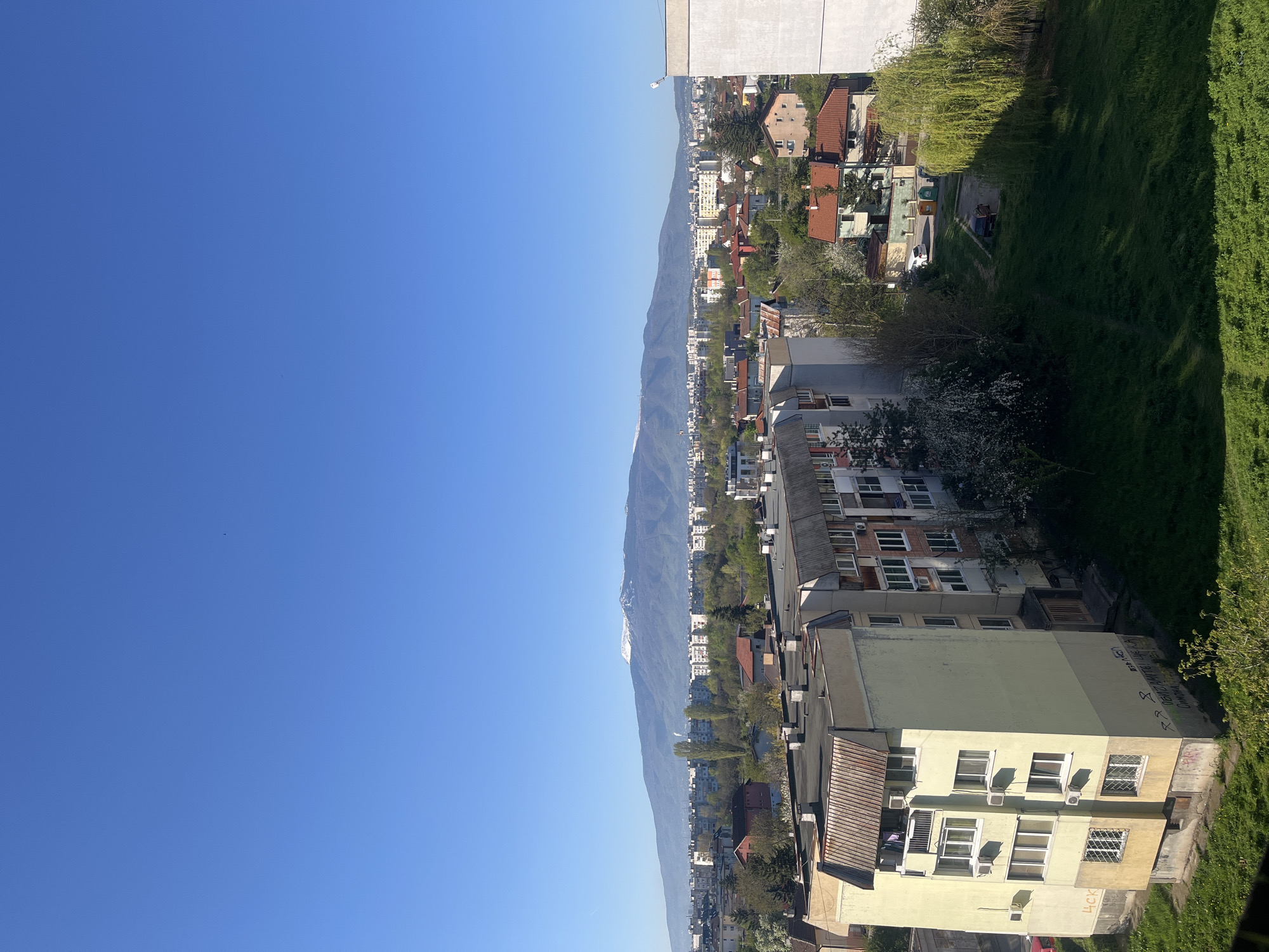 The image shows a view over Sofia, Bulgaria, from a high vantage point. The sky is clear and blue. In the distance is the Vitosha mountain, with a small amount of snow at its peak. The foreground shows rooftops of apartment buildings, some with air conditioning units. Greenery and trees are interspersed among the buildings, indicating the presence of parks or gardens. The scene is well-lit, suggesting a bright sunny day. The presence of residential buildings and the mountain backdrop suggest this is an urban environment within close proximity to nature.