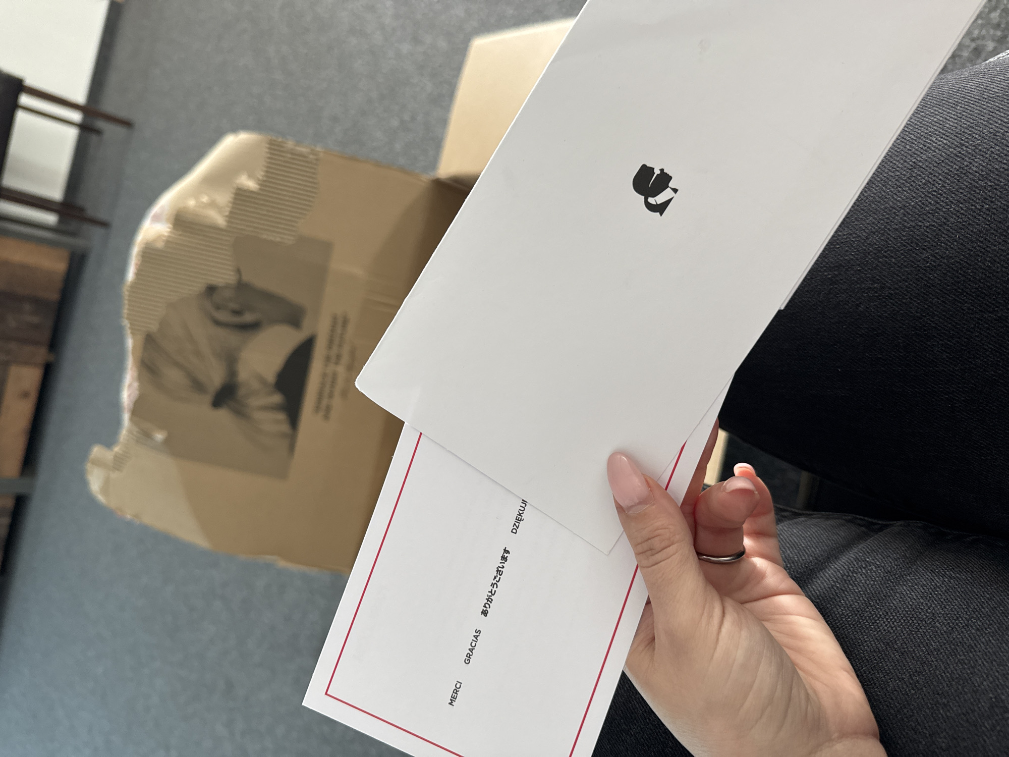 A high-angle, close-up shot captures a person's hand holding two cards over an opened cardboard box. The hand, adorned with light pink polished nails and a simple silver ring on the ring finger, holds a white envelope or card featuring a distinctive black, abstract logo that resembles two intertwined or stylized figures. Below this, a second white card with a thin red border is visible, displaying expressions of gratitude in several languages: "MERCI" (French), "GRACIAS" (Spanish), "ありがとうございます" (Japanese), and "DZIĘKUJĘ" (Polish).
To the left, an opened cardboard shipping box is partly visible, containing crumpled packing material and faint, illegible text or images on its interior. The person is wearing dark, possibly denim, pants, visible on the right side of the frame. The scene suggests an indoor setting, likely during the unboxing of a package. The lighting is even, without indicating a specific time of day. No emotions are explicitly visible, and there are no visual cues within the image itself to suggest the given location of Sofia, Bulgaria.