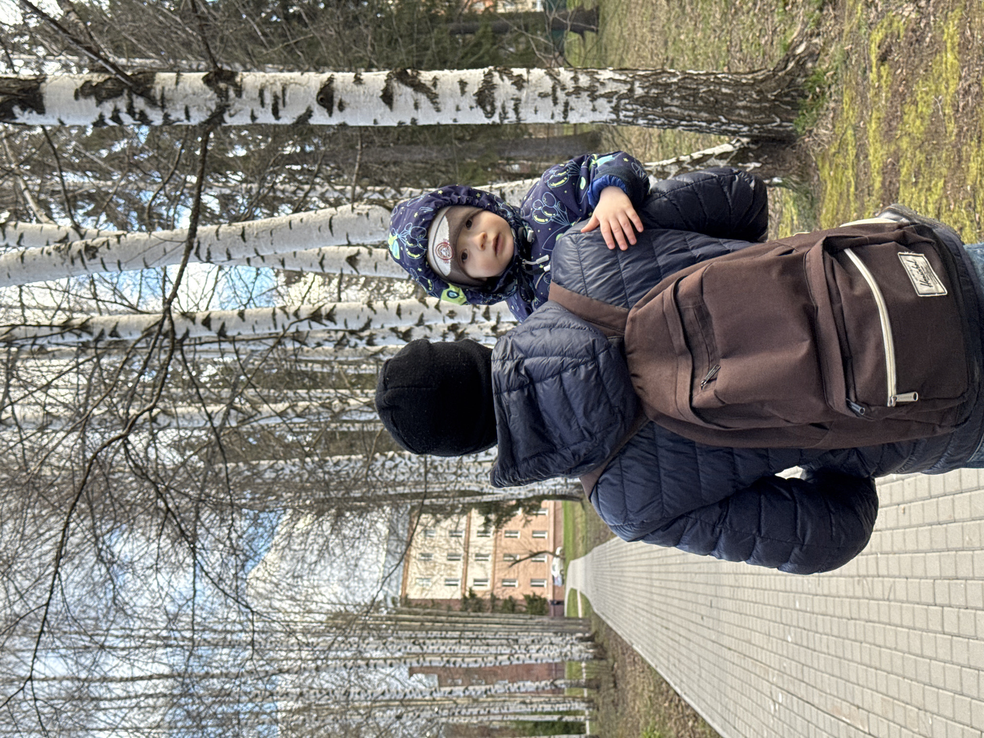 This image, rotated 90 degrees counter-clockwise, depicts a person carrying a young child on their back while walking outdoors. The carrier, seen from the back, wears a dark blue padded jacket, a black beanie, and blue jeans. A brown backpack with light-colored straps, featuring a "Herschel" brand label, is slung over the carrier's shoulders.
The child, a toddler, sits on the carrier's shoulders, looking towards the viewer with a calm expression. They are dressed in a dark blue hooded jacket adorned with a light blue pattern, and a light-colored beanie is visible under the hood.
The setting is an outdoor path, likely in a park or pedestrian area, lined predominantly with birch trees featuring their characteristic white and dark bark. Most trees are bare, suggesting a cooler season such as late autumn, winter, or early spring. The path itself is paved with light-colored square tiles. In the background, through the sparse tree branches, a multi-story building with a light brown/pink facade and multiple windows can be seen. The weather appears clear and bright during the daytime. The scene captures a casual outing in a location consistent with Izhevsk, Russia.