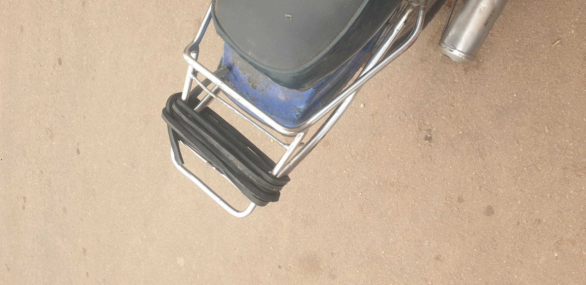 The image shows the rear of a motorcycle parked on a dirt road in Barakin Lerawi, Nigeria. The motorcycle has a black seat and a blue fender, with a chrome luggage rack on the back. Several black rubber straps are coiled on the luggage rack. A portion of the exhaust pipe is visible on the right side of the frame. The ground is a light brown, dusty dirt surface, with scattered small stones.