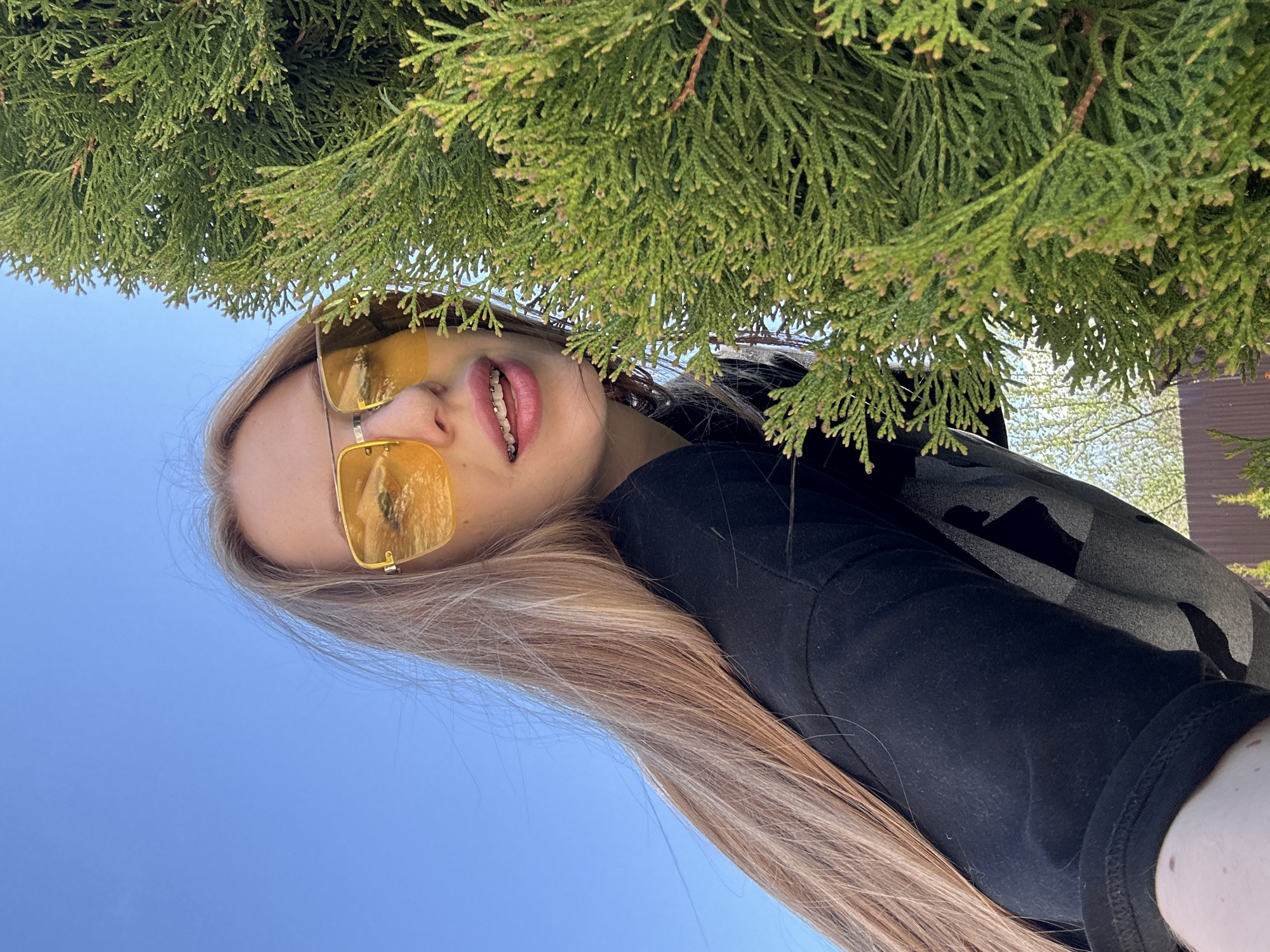 A young woman wearing gold-framed sunglasses is positioned beneath the foliage of a coniferous tree. She is wearing a black top with a patterned sweater. The background features a clear blue sky, suggesting an outdoor setting. The location is in the city of Chaikovsky, Russia. The image is taken from a low angle, looking up towards the woman. The woman's face is visible, including her braces.