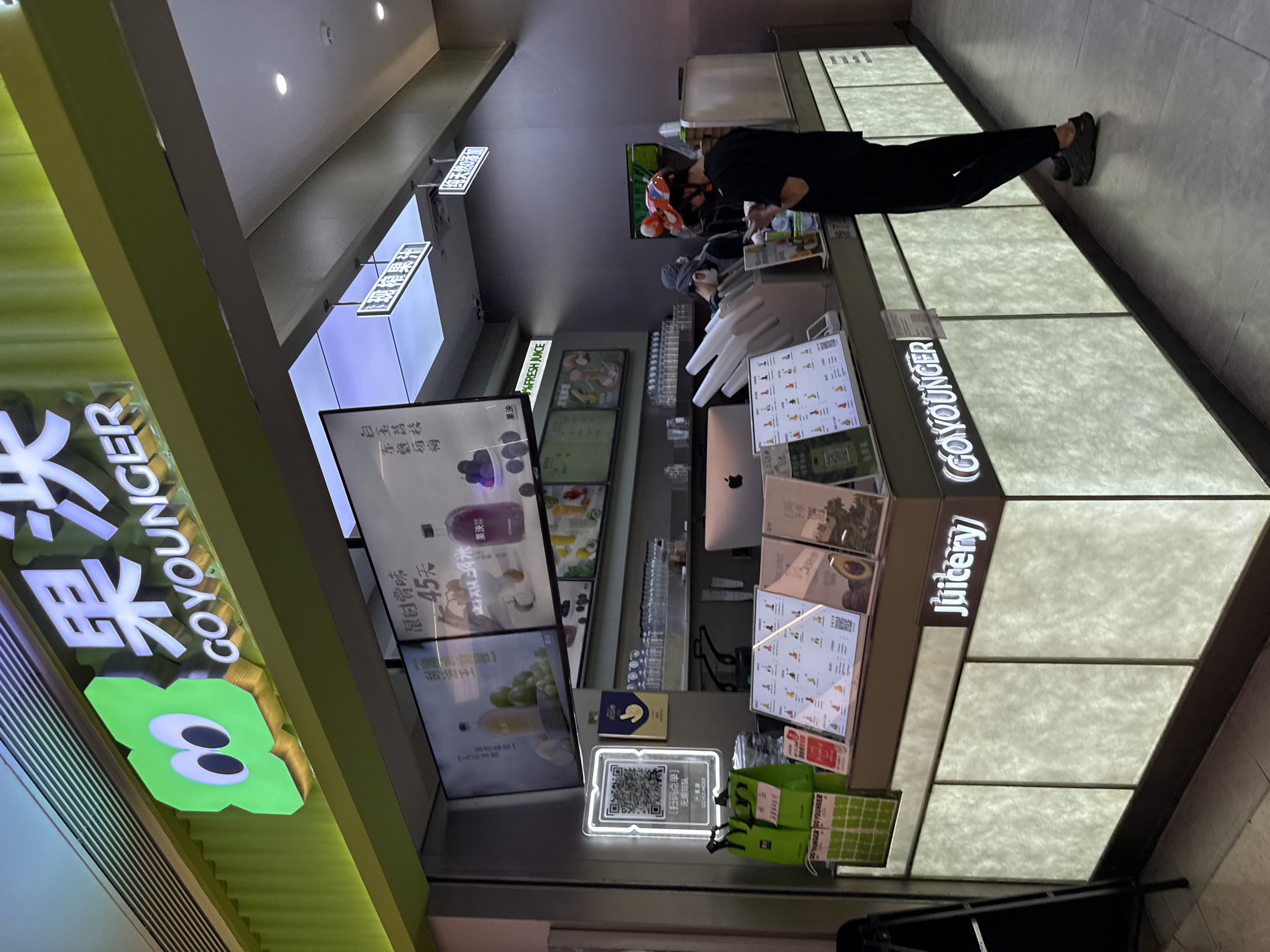 The image shows a juice bar named "GO YOUNGER" located in the Haizhu District, China. The exterior of the juice bar is brightly lit with a prominent green logo featuring two large eyes. The interior displays are digital menus showcasing various fruit juices and prices, with one menu item priced at 45 yuan. There are several green shopping bags stacked near the counter, and a QR code is displayed for ordering. Two employees are visible behind the counter, one preparing drinks and another interacting with a customer. A customer, wearing a colorful helmet, is standing at the counter. The juice bar counter itself is illuminated with a soft, ambient light and features the "Juicery" logo. The overall environment appears to be a modern, clean, and inviting retail space.
