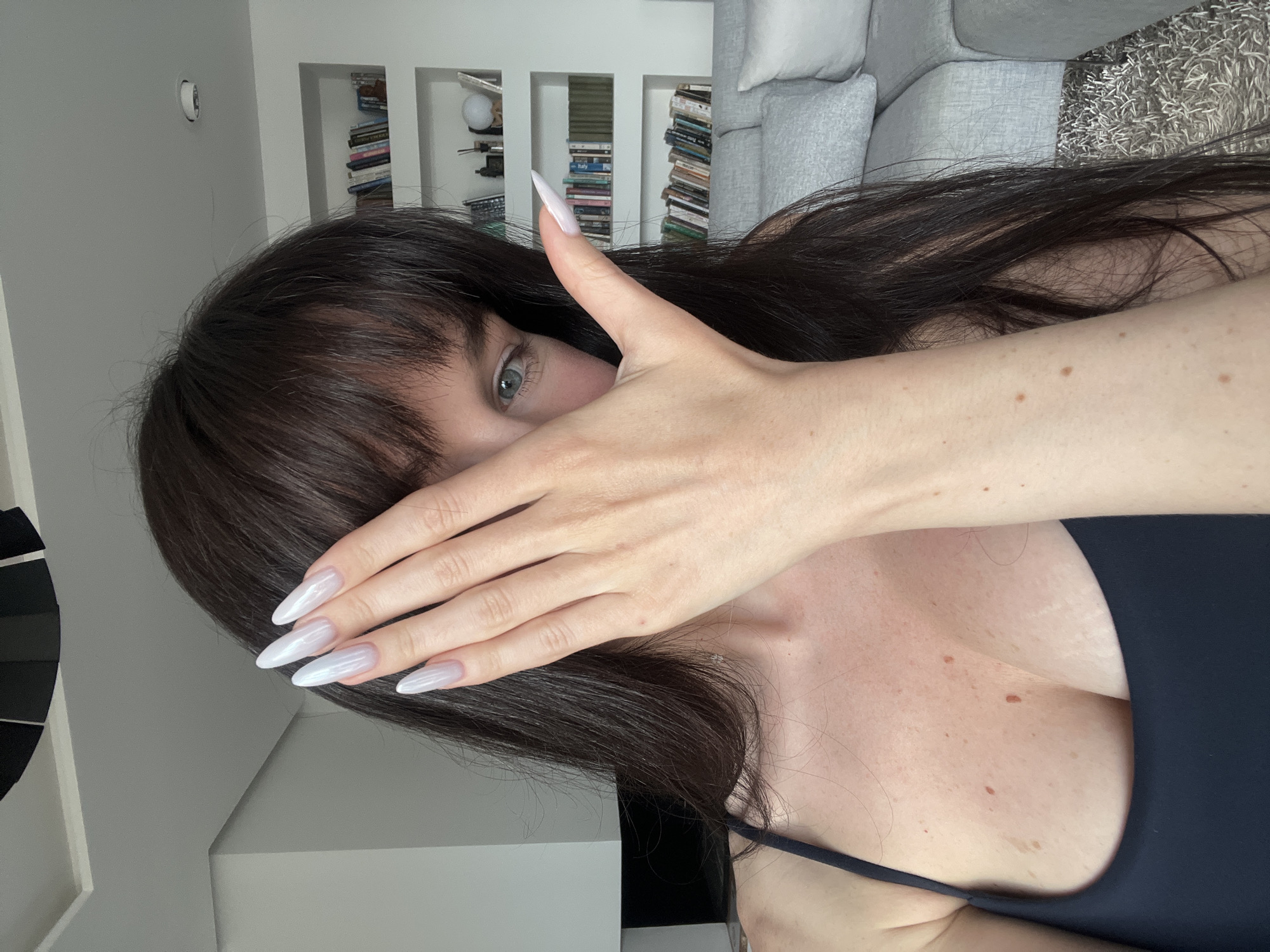 A woman is pictured indoors, partially obscuring her face with her right hand. Her long, dark hair with bangs frames her face. Her eye is partially visible. She wears a dark-colored garment with a thin strap. Her skin is pale with some freckles. Her fingernails are long, pointed, and white with a pearlescent sheen. A bookshelf filled with books and a gray sofa are visible in the background. The location in the city of Ivanovići, Montenegro, cannot be determined based on the provided image.