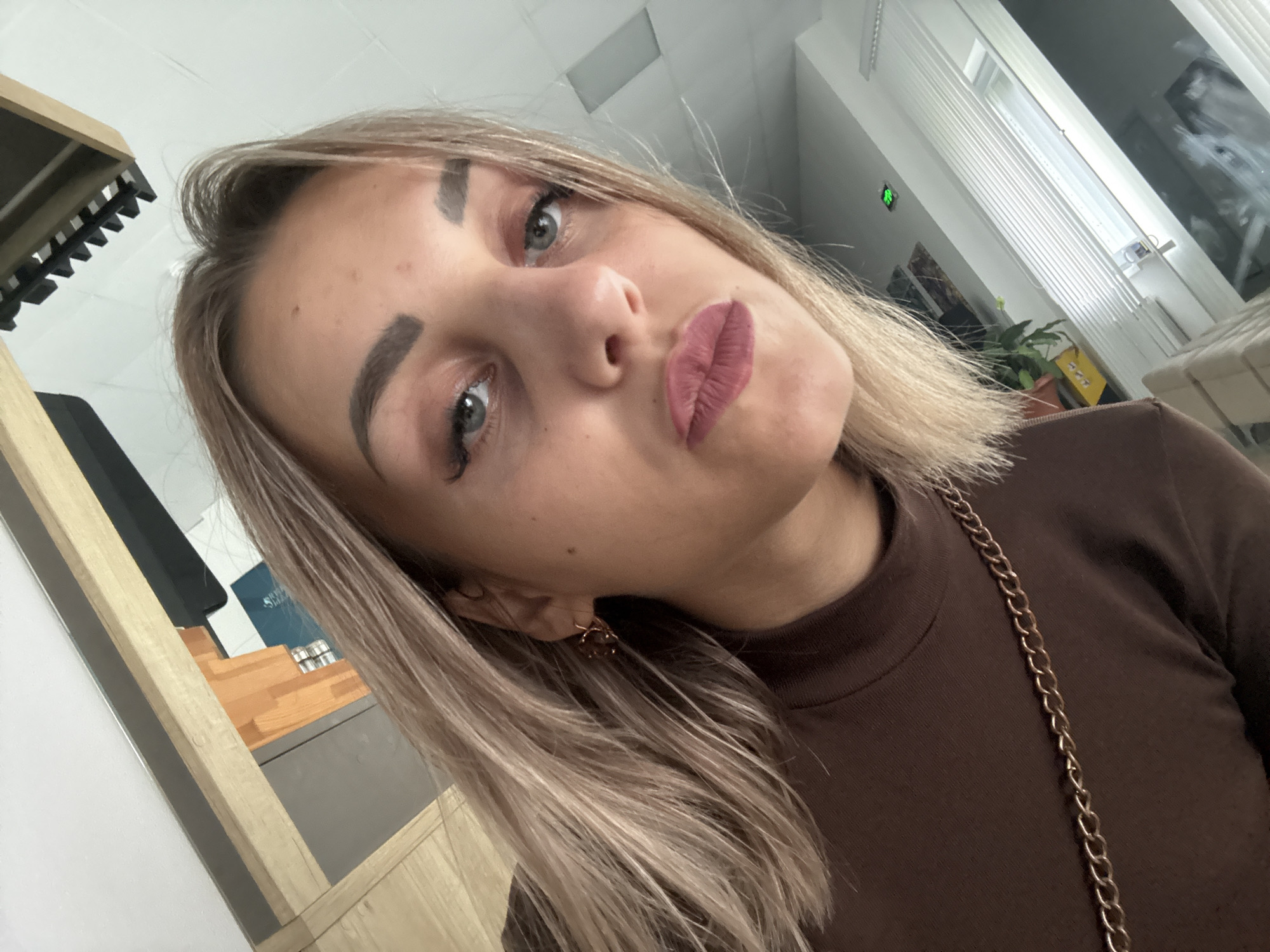 A woman with blonde hair and visible makeup is looking directly at the camera. She is wearing a brown turtleneck shirt with a gold chain necklace. The background suggests an indoor environment, possibly an office or room, with some shelving and blinds visible. The lighting is bright, and the image appears to be a selfie. There are no discernible indications of the specific location within Yuzhno-Sakhalinsk, Russia.