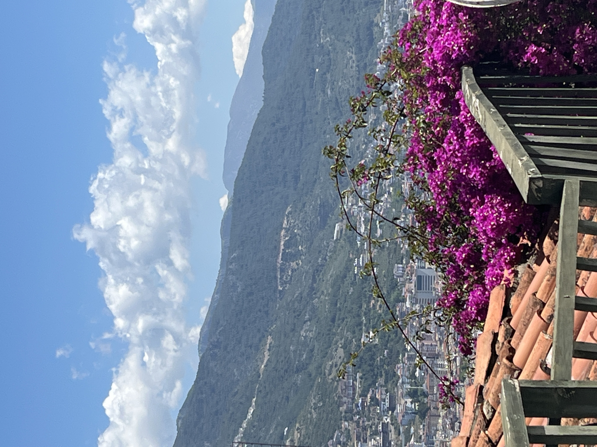 The image depicts a landscape scene in Alanya, Turkey. The foreground features a red-tiled roof with a weathered wooden railing. Bright pink bougainvillea flowers cascade over the railing and roof tiles.
In the midground, a densely populated area is visible, with numerous buildings and structures scattered across a hillside that slopes downwards. A modern skyscraper is visible, marking a location in the city of Alanya.
The background shows a mountain range that recedes into the distance, with varying shades of green vegetation covering its slopes. The sky is predominantly blue, with patches of white, fluffy clouds.
