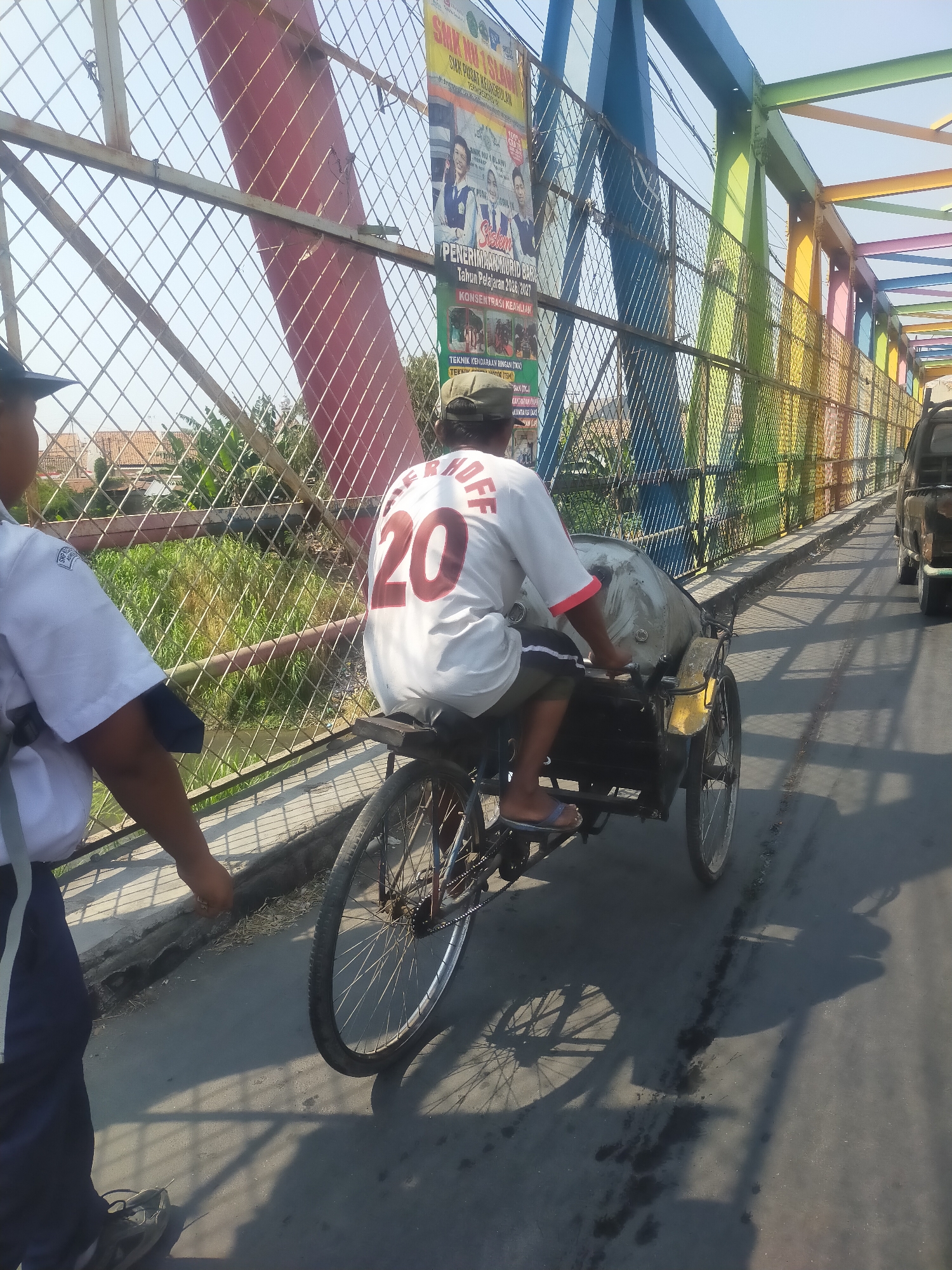 An outdoor, eye-level, medium shot captures a colorful bridge in Tembok Banjaran, Indonesia, on a sunny day. The bridge's metal structure is painted in vibrant hues of red, blue, green, yellow, pink, and orange. A metal mesh fence runs along the side of the bridge, with posters advertising an educational institution on the left side. In the foreground, a person wearing a white uniform with dark blue accents, likely a student or staff member, walks towards the left edge of the frame. To the right of the uniformed person, a man is riding a three-wheeled bicycle rickshaw, also known as a becak. The becak is yellow and black and has a large, covered passenger compartment behind the rider. The man is wearing a white t-shirt with the number "20" in red on the back and dark shorts. He has sandals on his feet. The becak is moving forward on the bridge, casting a shadow on the asphalt road. In the background, lush green vegetation and a few houses are visible below the bridge, suggesting a semi-rural or suburban setting. A dark-colored truck is also visible further down the bridge. The scene depicts a typical day on a local bridge, with everyday transportation and urban infrastructure.