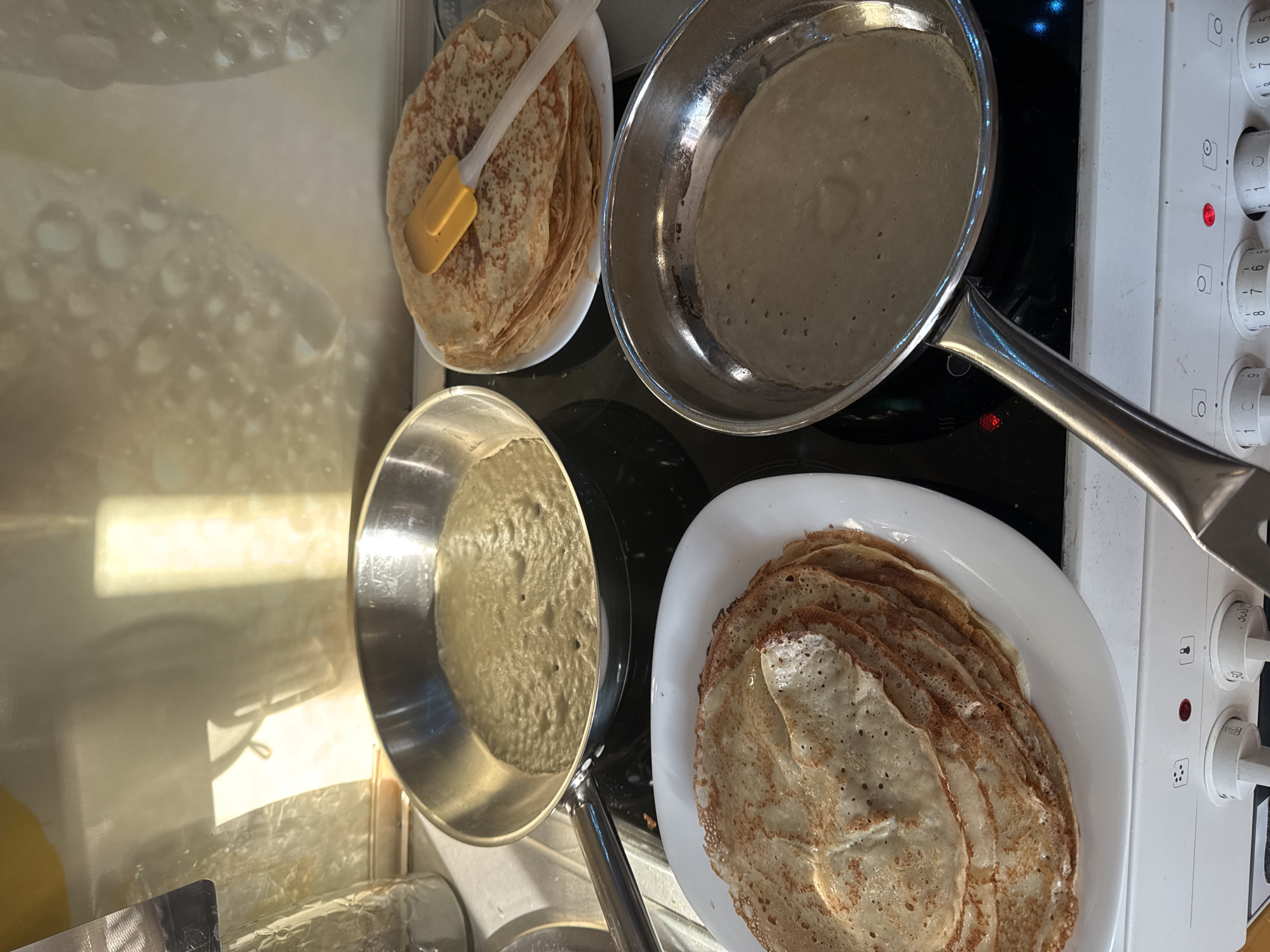 The image presents an overhead view of a kitchen countertop and an electric stovetop, depicting the ongoing process of making pancakes or crepes.
In the upper right, a stainless steel frying pan holds a light-colored batter, showing some bubbles, indicating it is actively cooking on a burner. Next to it, to the left, a white plate is laden with a stack of golden-brown, cooked pancakes, and a yellow-bladed spatula rests on top of the pile.
Another stainless steel frying pan, also containing pancake batter, is positioned on a burner in the lower left portion of the image. Adjacent to this pan, at the bottom right, a large stack of cooked pancakes is neatly arranged on a white oval or rectangular serving plate.
The right side of the frame features the white control panel of an electric stove, equipped with several rotary knobs marked with numerical settings (e.g., 0-9, 1-6) and universal symbols for different cooking functions. Red indicator lights are illuminated, suggesting that at least one burner is active.
Bright reflections, possibly from a window or overhead light source, are visible on a reflective surface in the upper left and central left areas, hinting at a brightly lit kitchen. The scene captures the industrious atmosphere of preparing a significant quantity of pancakes.