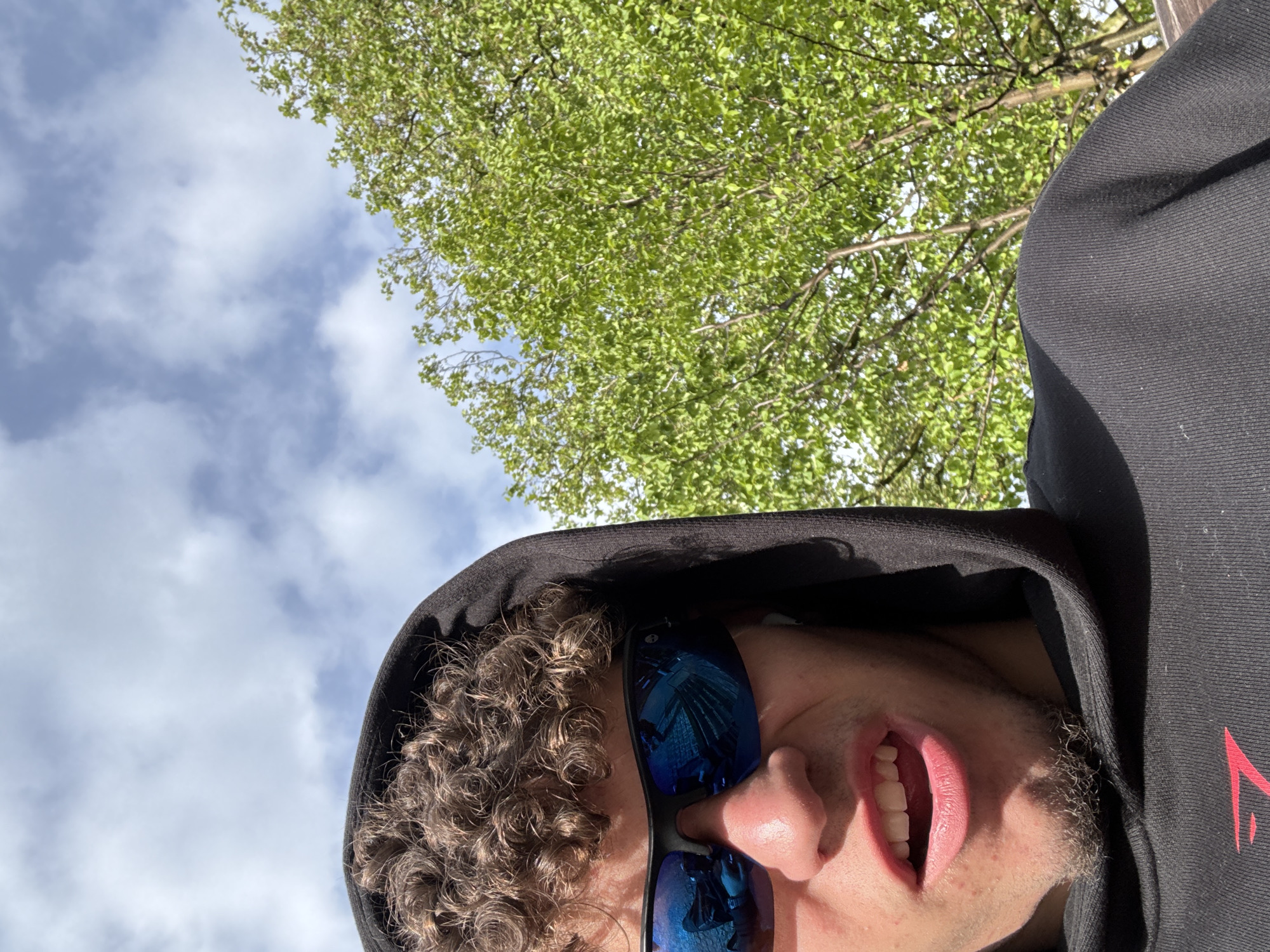 The image is a selfie taken from a low angle, looking up. A person with curly brown hair, wearing a black hooded sweatshirt and blue mirrored sunglasses, occupies the lower right quadrant. The person's mouth is open. The background shows a cloudy sky and tree branches with green leaves. Based on the reflected buildings in the glasses, I have no enough data to infer the location in the city of Aachen, Germany.