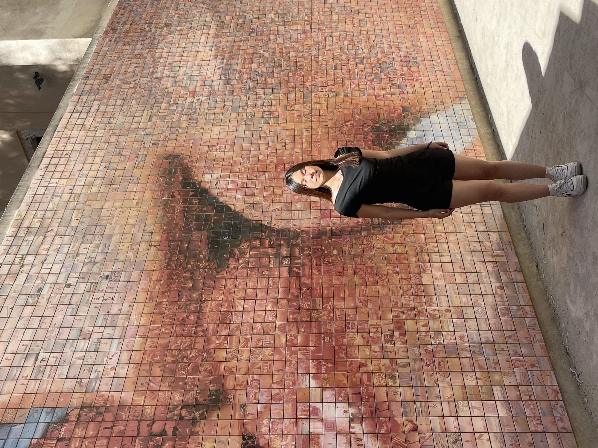 In Barcelona, Spain, a young woman stands in front of a large, tiled mural. The mural is comprised of many small squares, each depicting an image, possibly photographs or painted tiles. The overall effect of the mural appears to be a larger image, with a dominant reddish-orange hue. The woman is wearing a black, off-the-shoulder top and black shorts, paired with white sneakers. Her eyes are closed, and the sunlight is casting a shadow on her face. She is standing on a textured ground, and a shadow of an oval shape is cast on the ground to her right, suggesting the presence of another object or structure not fully visible in the frame. The background includes the continuation of the tiled wall and a hint of a building above.