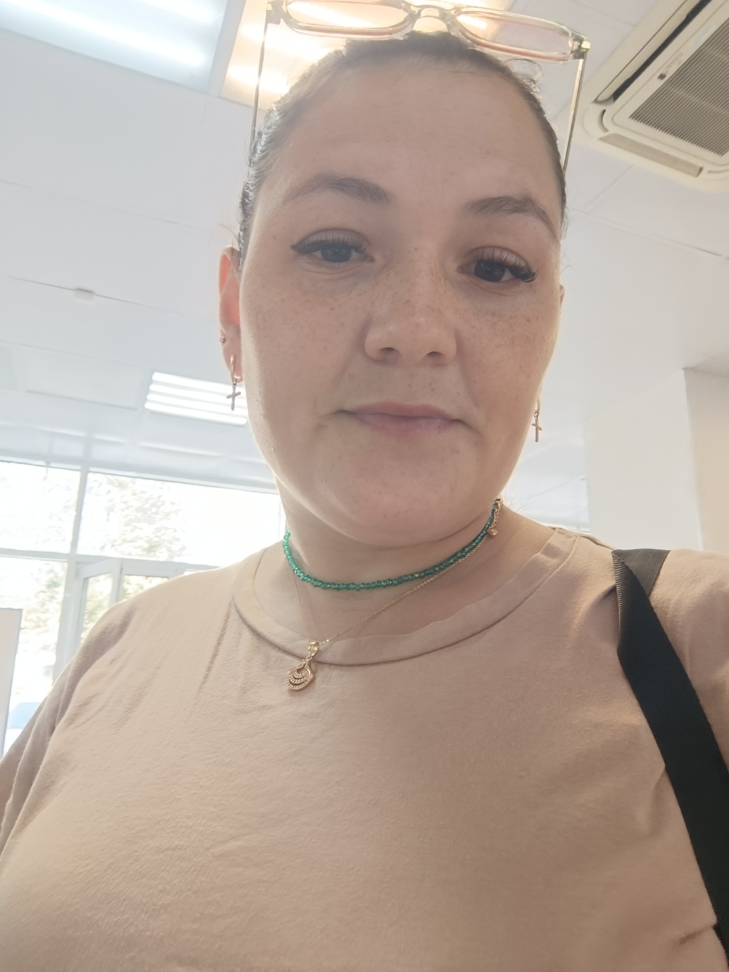 A woman is taking a selfie indoors, likely in an office or commercial space. She has fair skin with freckles, dark hair pulled back, and is wearing glasses on top of her head. She has on a tan t-shirt, a green beaded necklace with a gold pendant, and small gold cross earrings. In the background, there are fluorescent lights, a ceiling air conditioning unit, and large windows with a view of trees and buildings, suggesting an urban or suburban environment. The woman appears to be alone and is looking directly at the camera.
